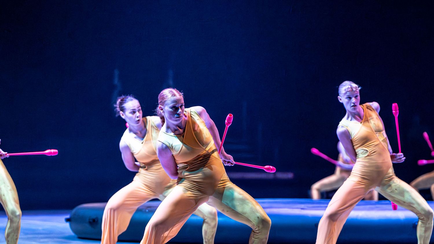 Female gymnasts in nude costumes perform with pink clubs under blue stage lighting.