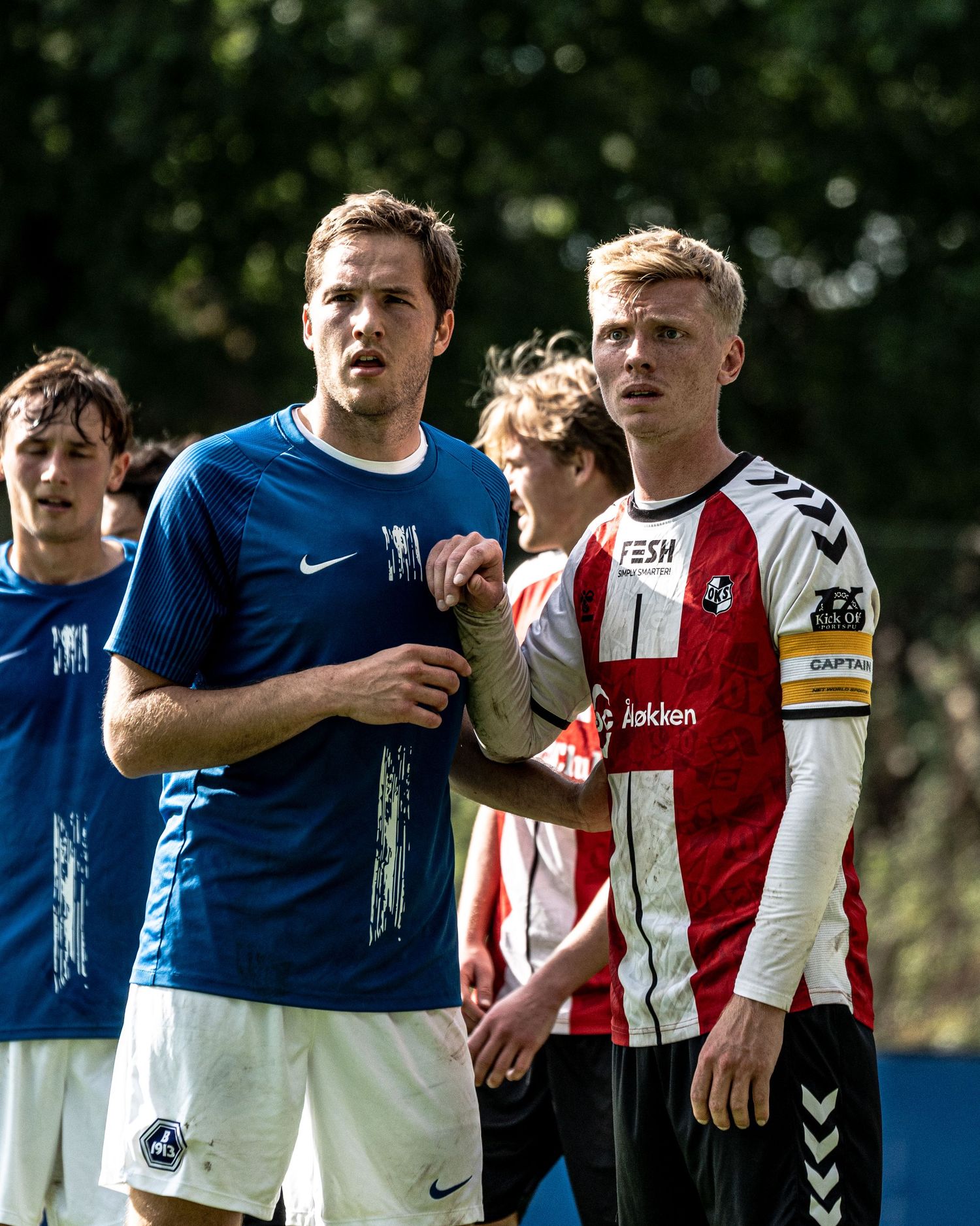 Two soccer players, one in blue and one in red and white Hummel kit, stand together during a match.