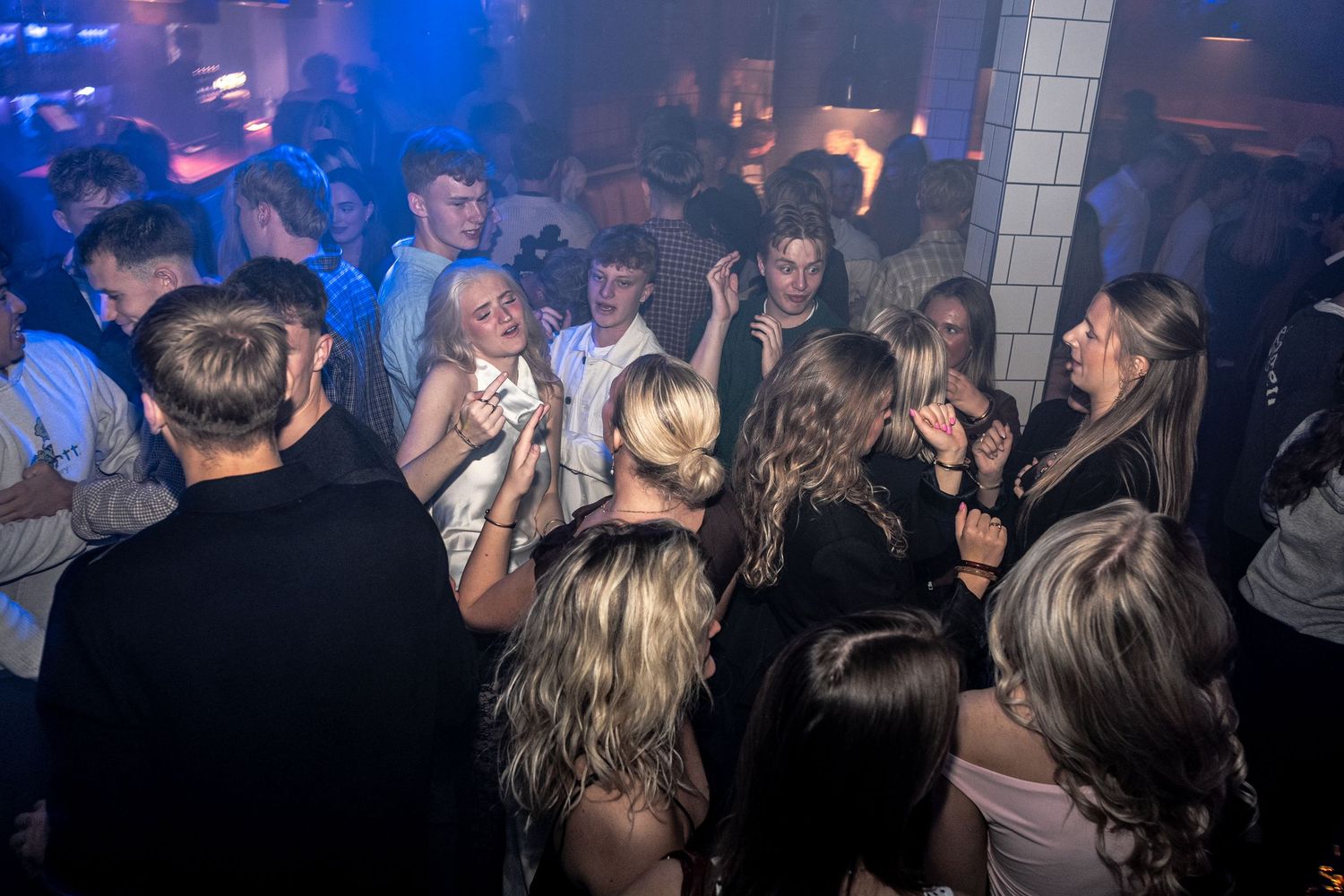 Crowd of young partygoers dancing and laughing together in a busy nightclub with blue lighting.