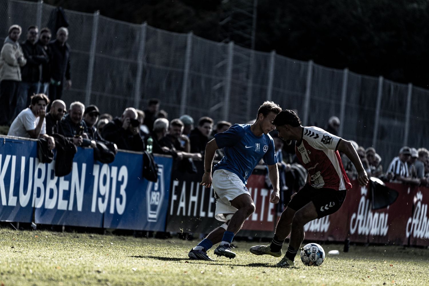 Two soccer players battle for the ball near the sideline in front of a crowd.