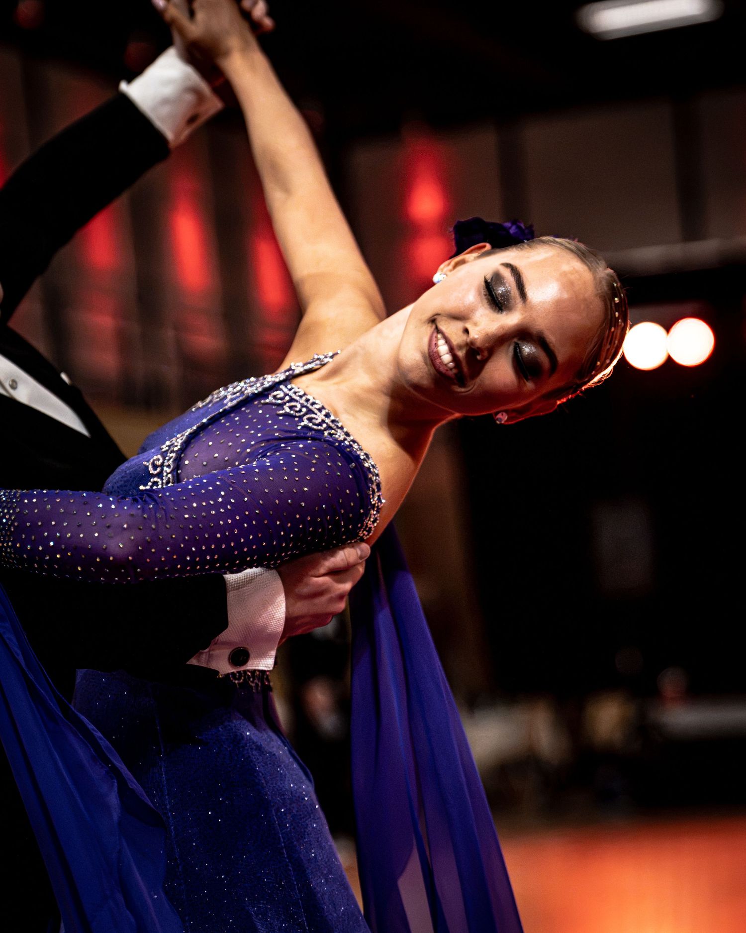 Female ballroom dancer in blue rhinestone costume smiles while performing elegant dip with partner on competition floor.