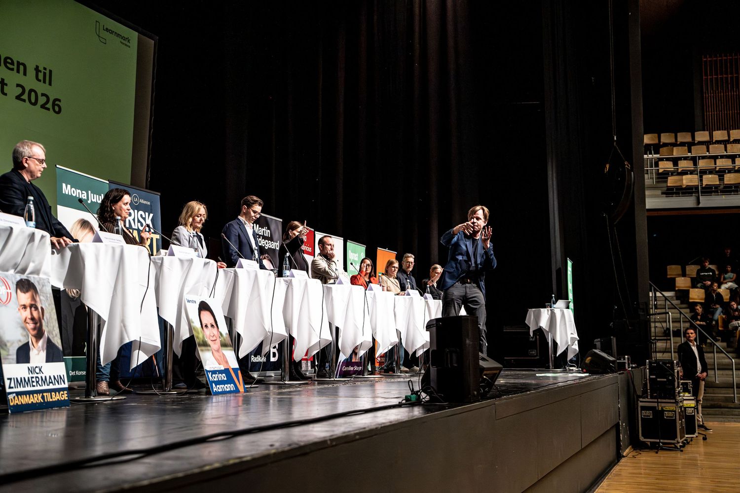 Large panel debate on stage with multiple participants seated at white draped tables.