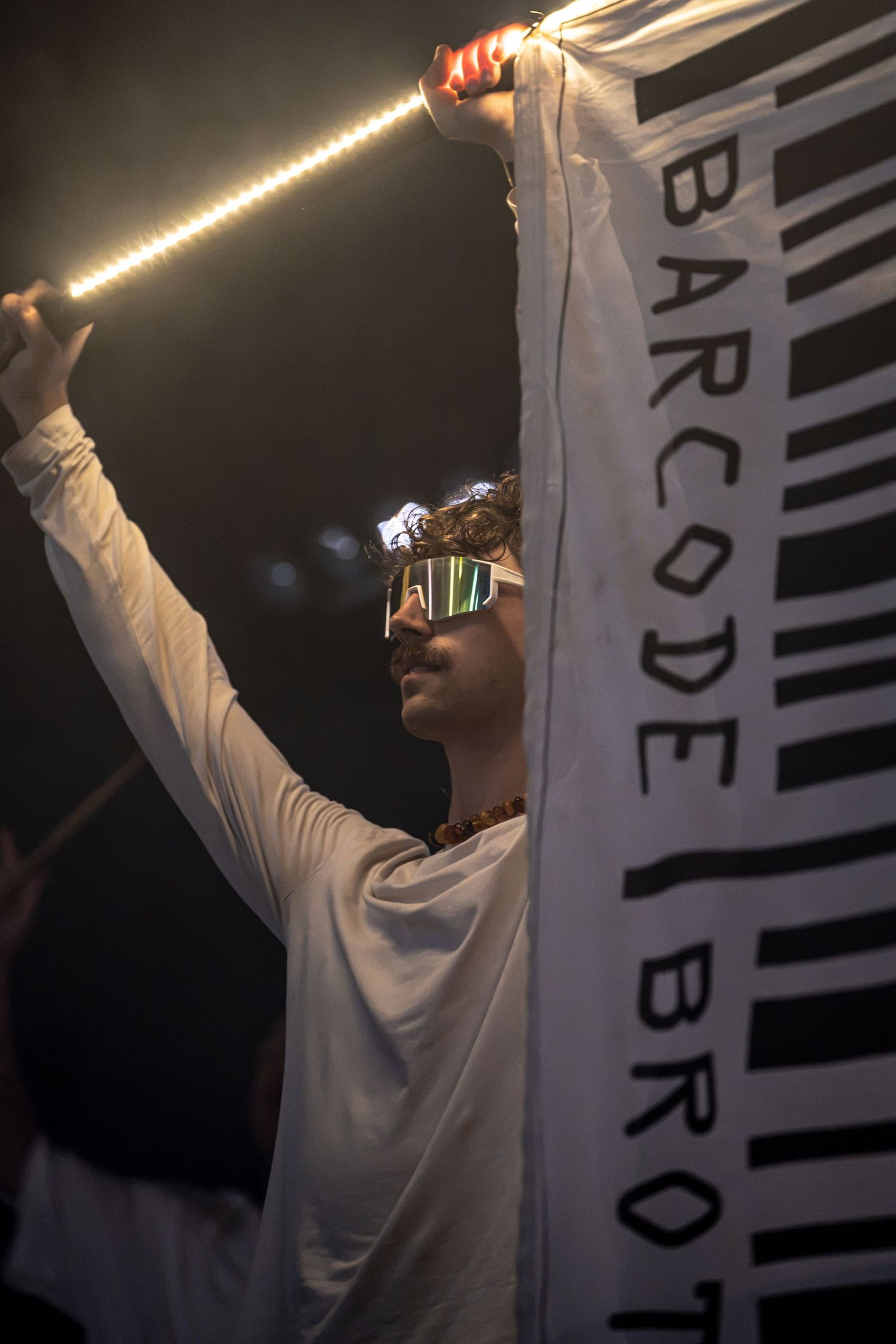 Performer in white outfit holds LED light bar and Barcode Brothers flag at live show.