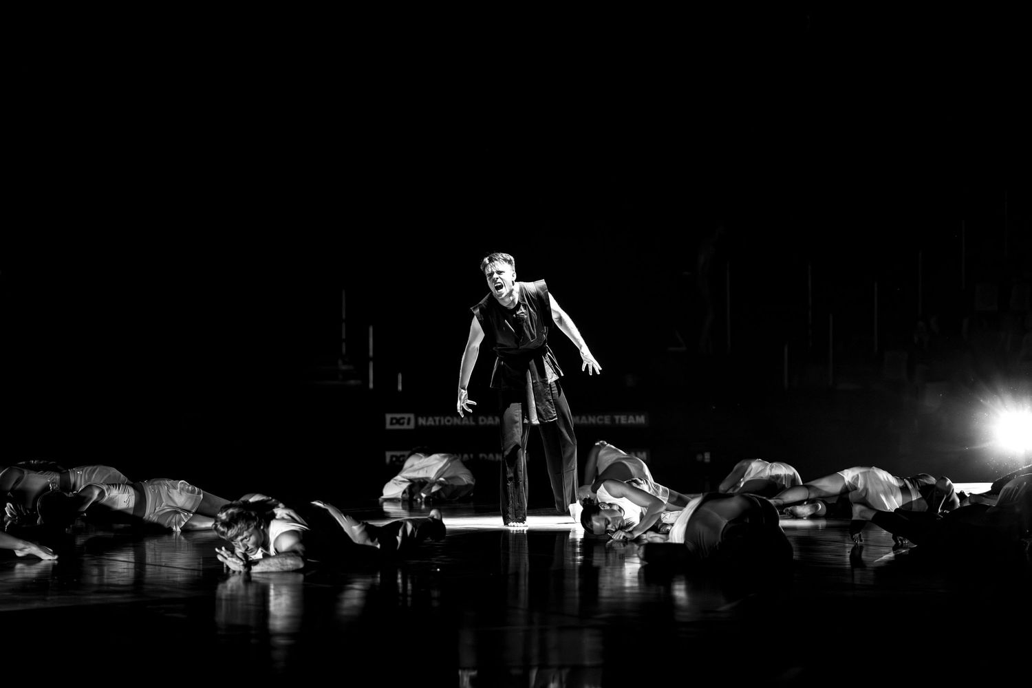 Black and white photo of solo dancer standing on stage surrounded by performers on floor.