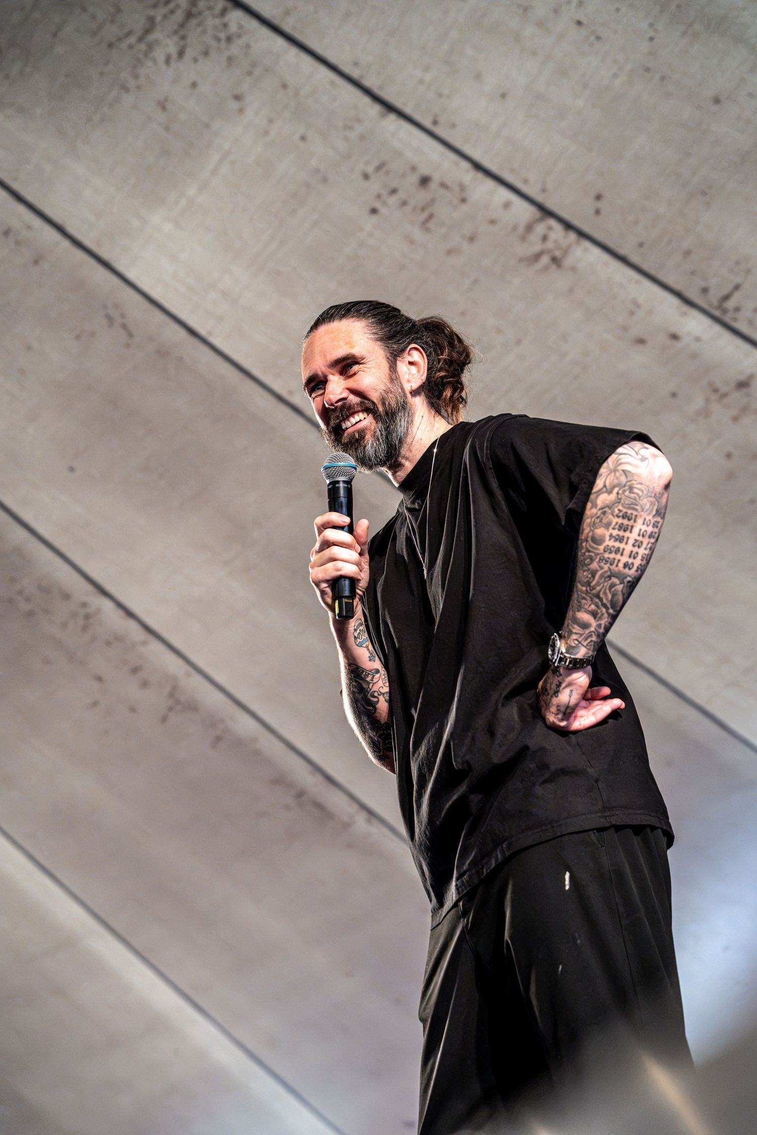 Smiling tattooed bearded performer with hair bun bowing on stage while holding microphone in black outfit.