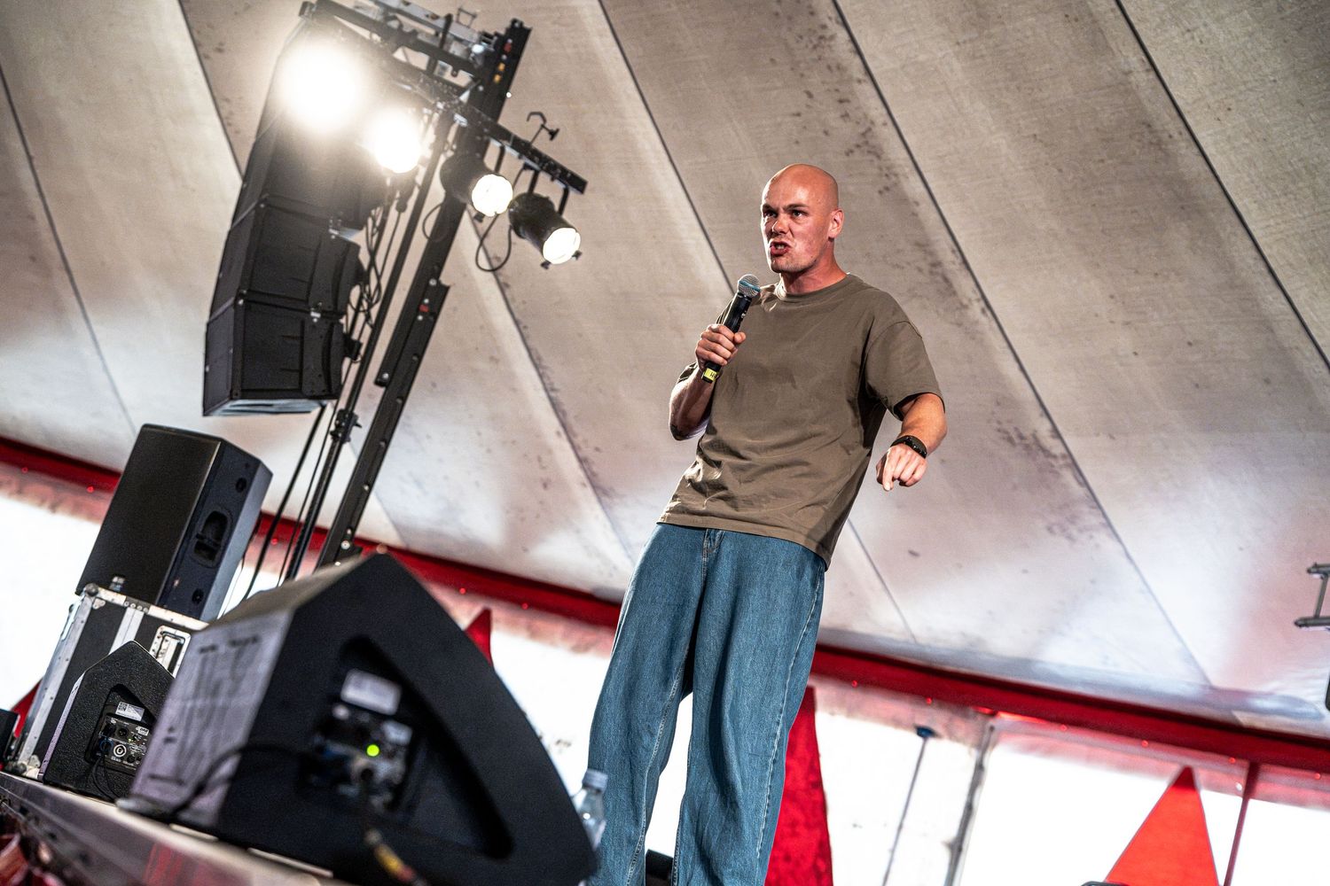 Bald male performer speaks into a microphone on an outdoor festival stage under a tent canopy.