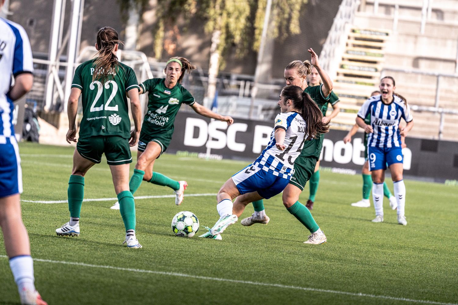 Female soccer player #17 in blue and white strikes the ball surrounded by green-uniformed opponents.