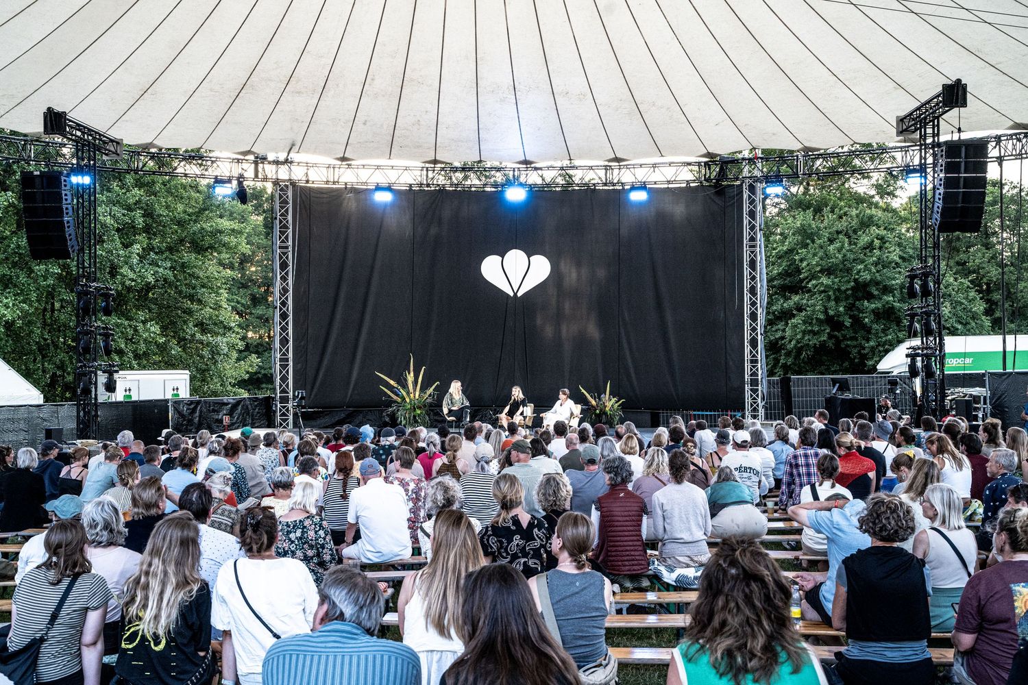 Large crowd gathered at outdoor stage event with heart logo backdrop.