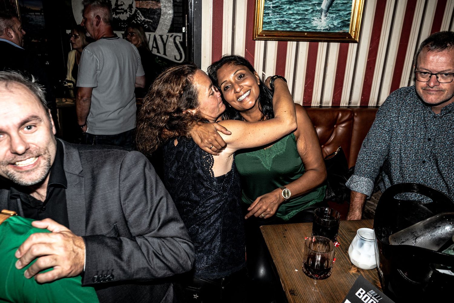 Two women embrace joyfully in a lively pub booth while friends smile and chat nearby at a wooden table with drinks.