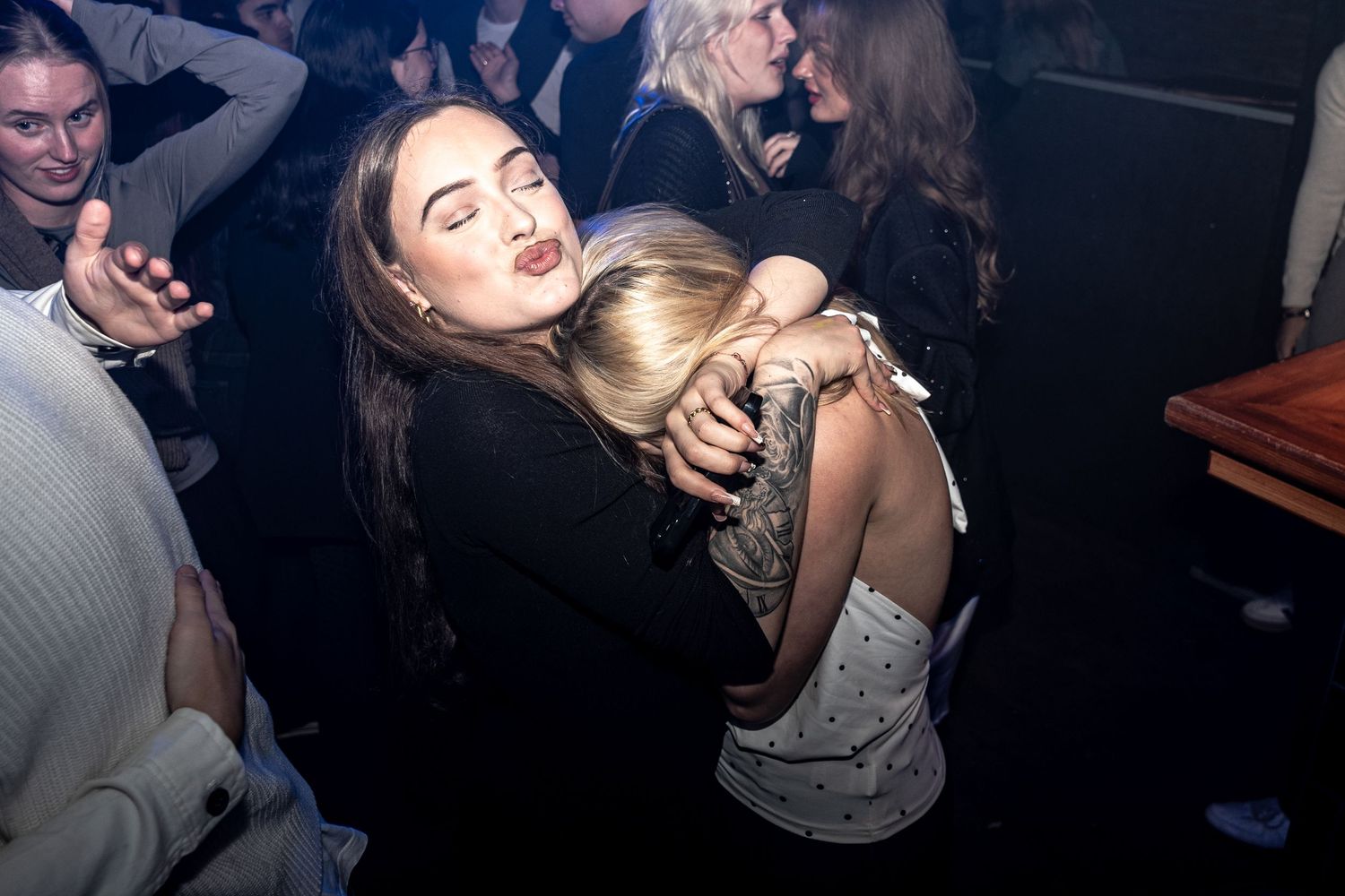 Two women embrace on a crowded nightclub dance floor while friends laugh and celebrate in the dark background.