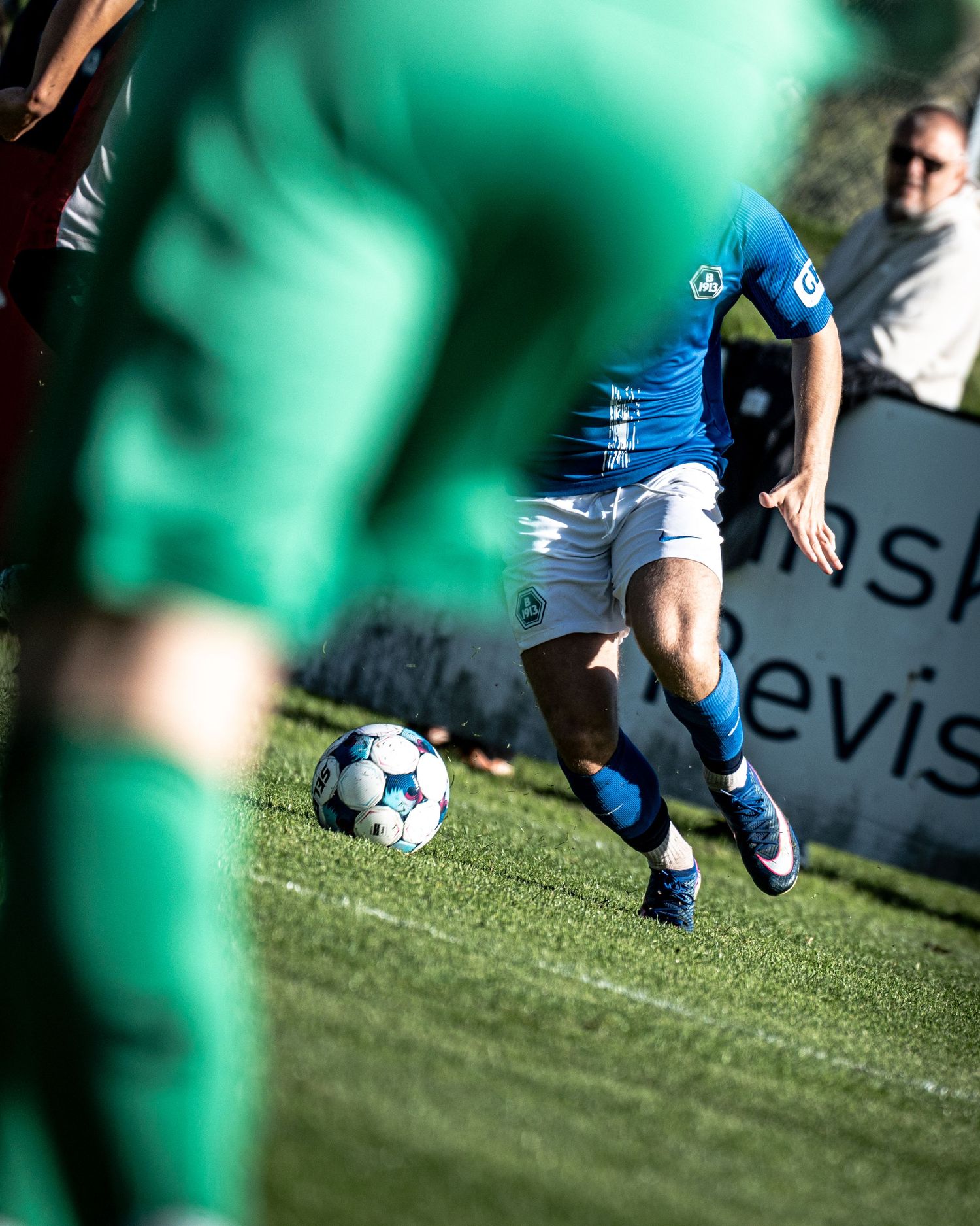Soccer player in blue and white kit dribbles ball past green-shirted opponents on grass.