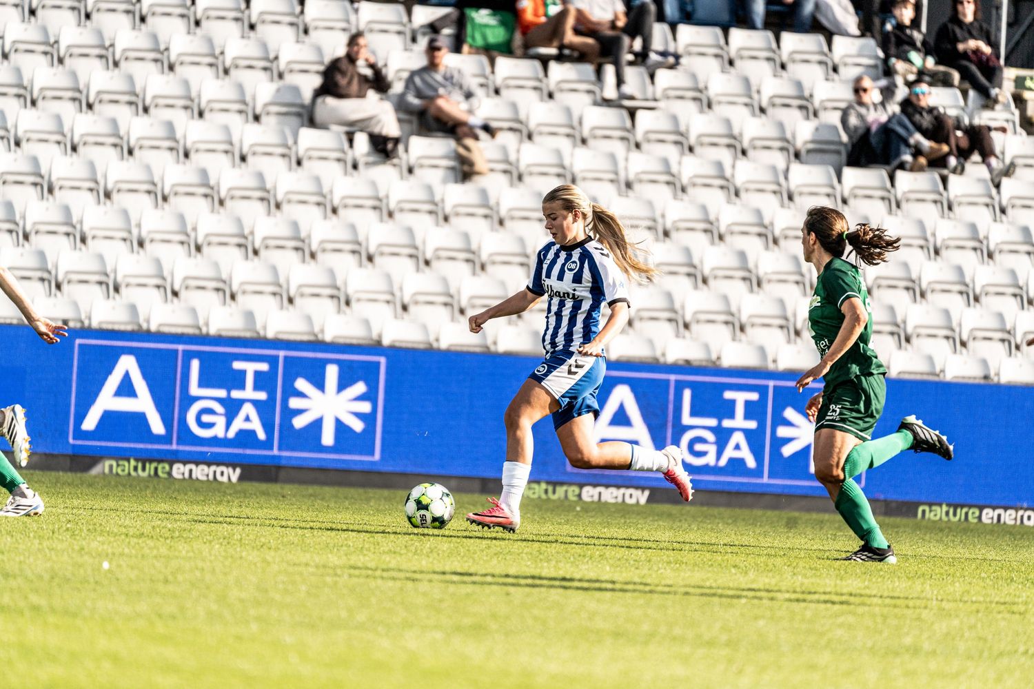 Female soccer player in blue and white kit passing ball during a sunny Liga league match in stadium.