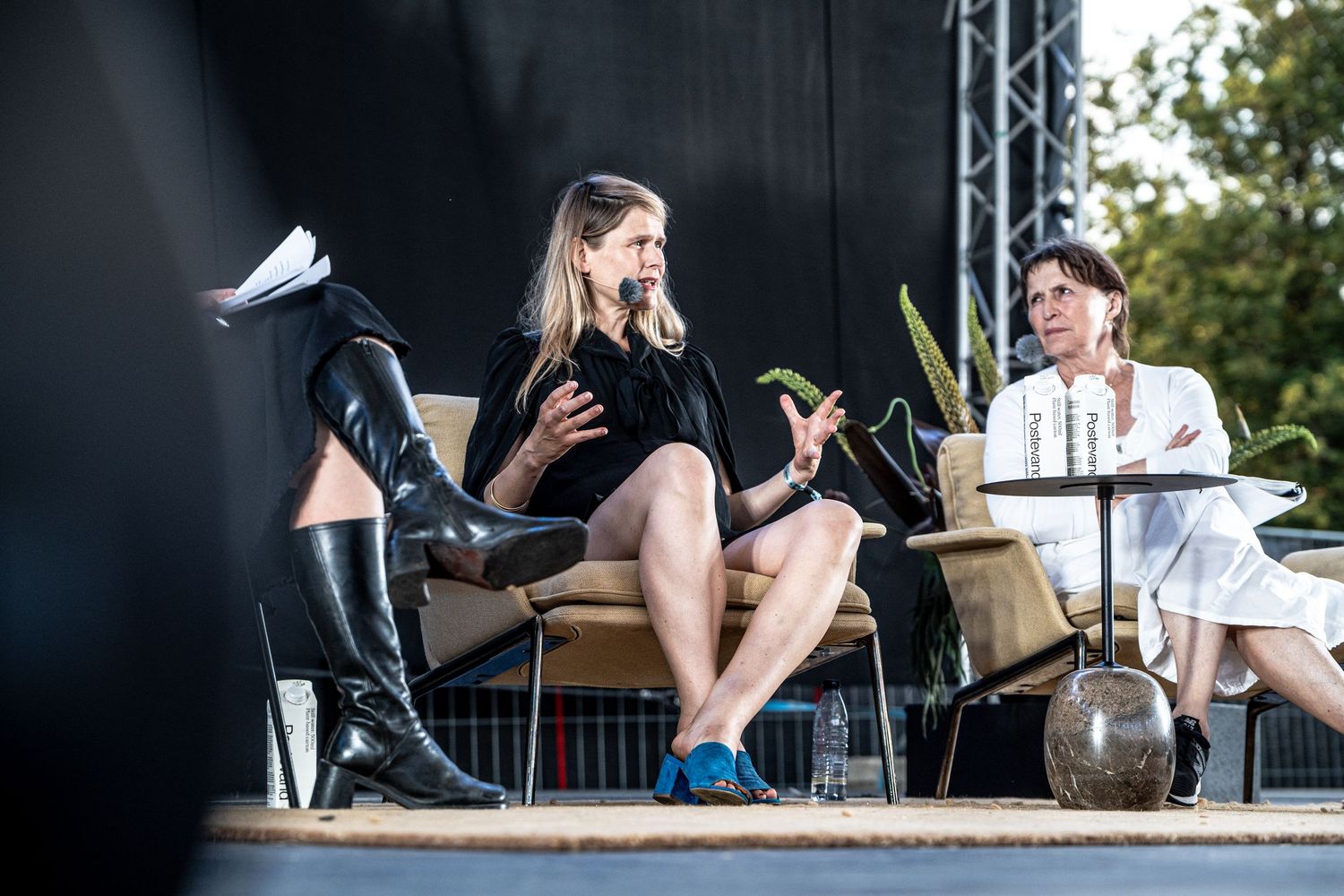 Woman in black dress speaking and gesturing during an outdoor stage panel discussion with plants nearby.