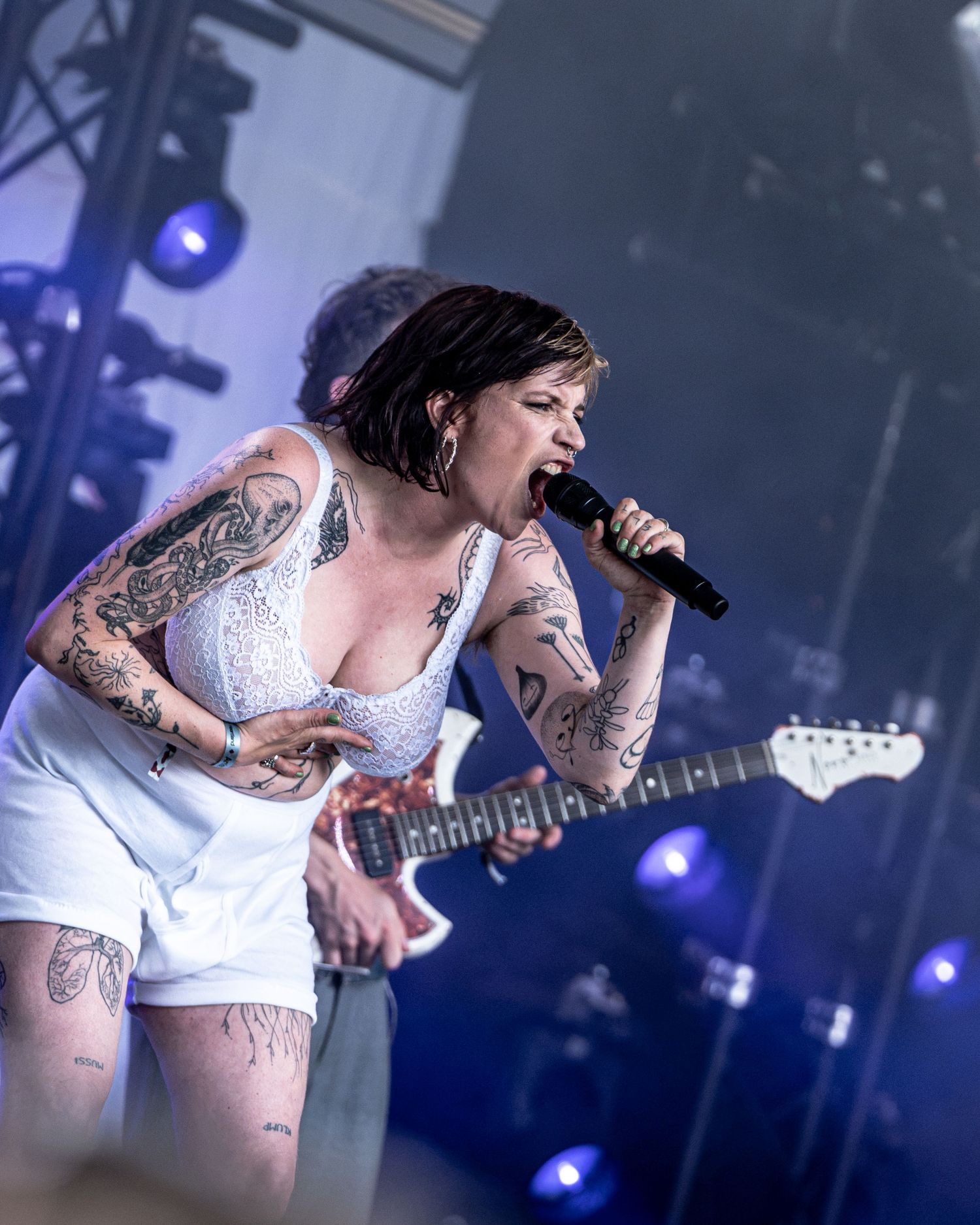 Tattooed female guitarist sings into microphone wearing white outfit on outdoor stage.