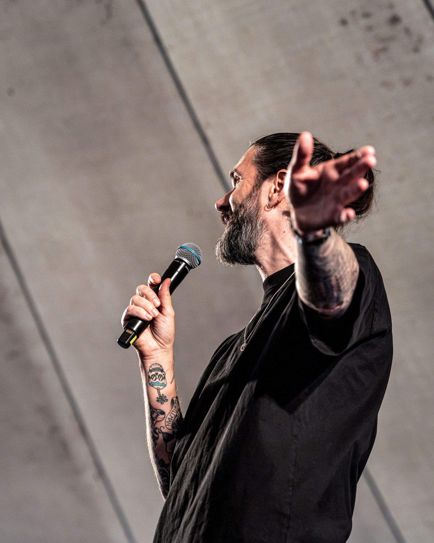 Bearded tattooed male singer in black shirt performing on stage holding microphone with arm outstretched.