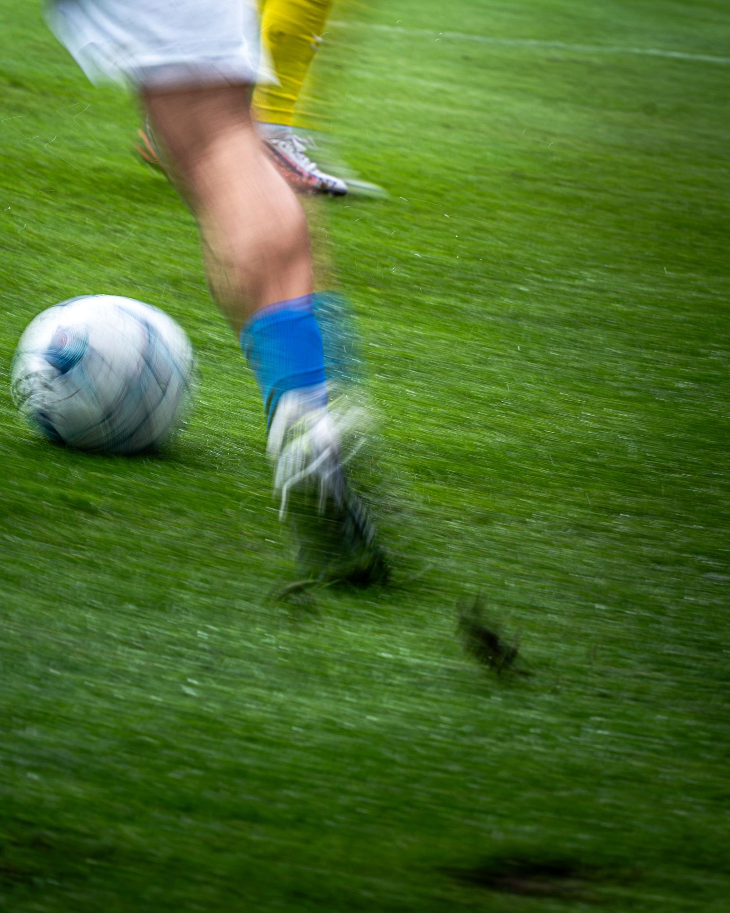 Soccer player in motion kicking a white ball on green grass, wearing blue socks and cleats, with motion blur effect.