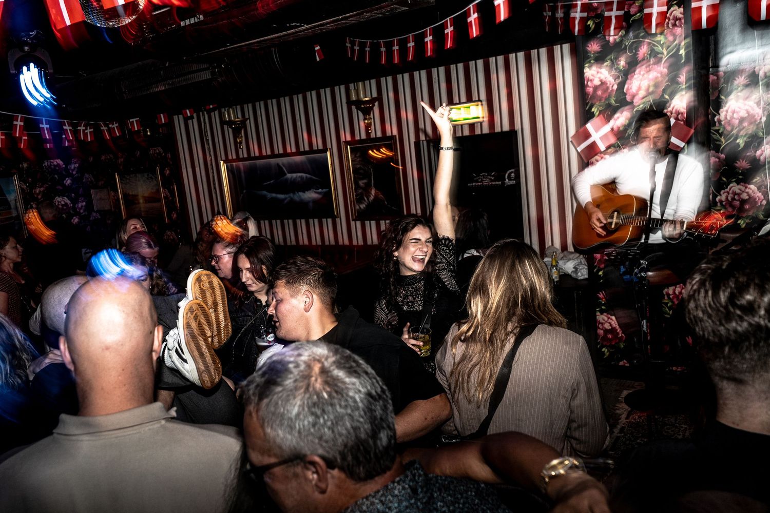 Crowded bar with a cheering woman raising her arm as a guitarist plays live music beneath red-and-white striped decorations.