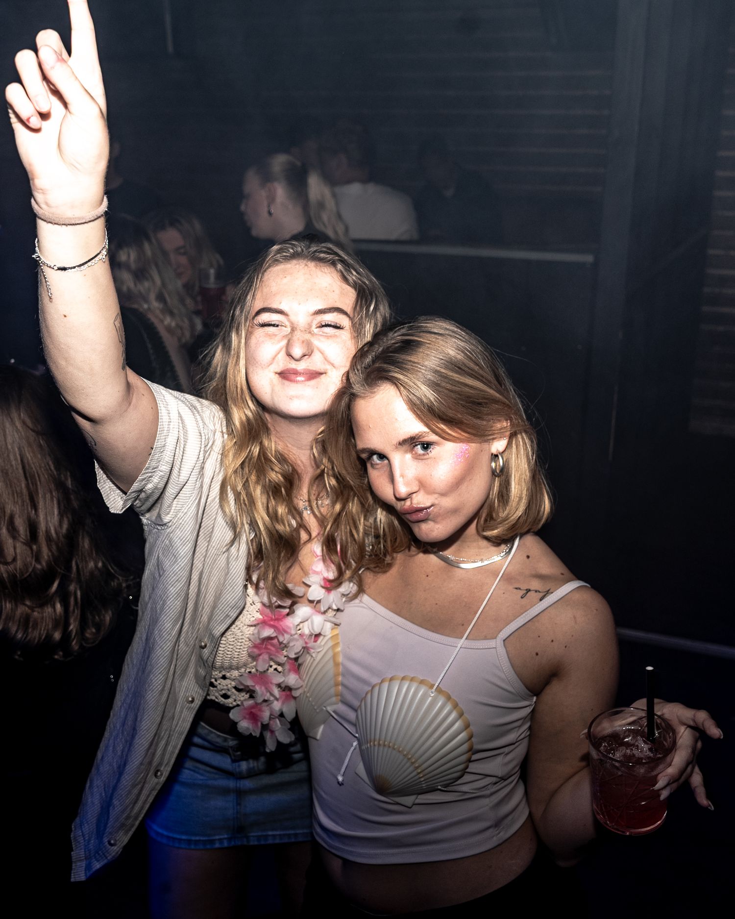 Two smiling young women pose together at a nightclub, one raising her arm in the air and the other wearing a seashell top and holding a red 