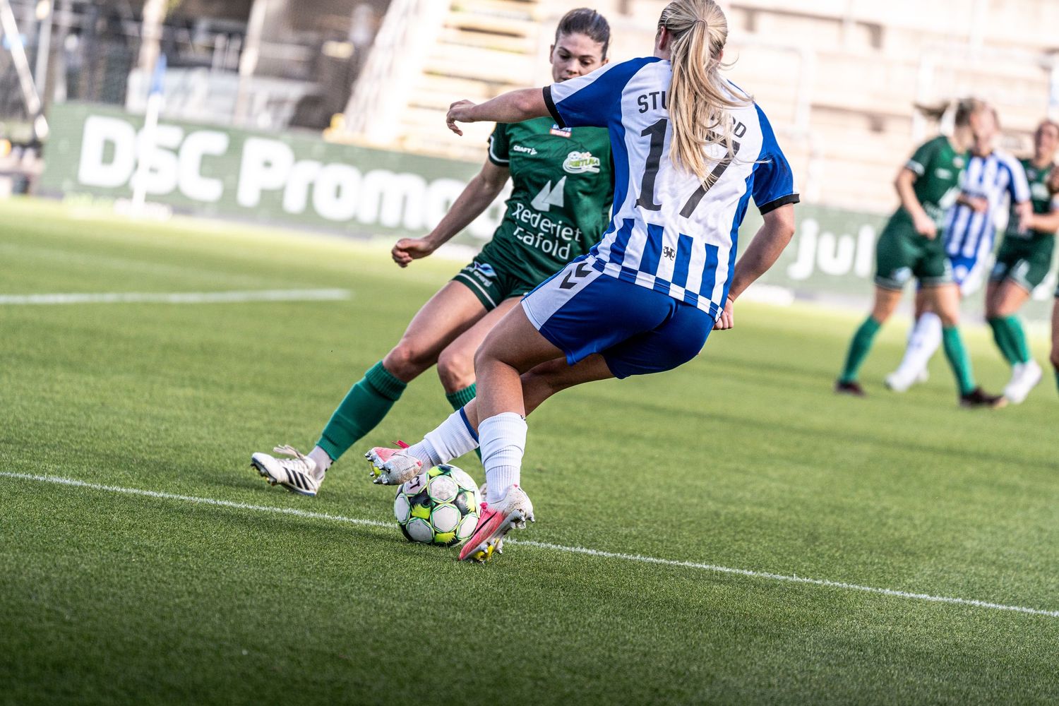 Female footballers in green and blue-white kits battle for possession during a women's soccer match.