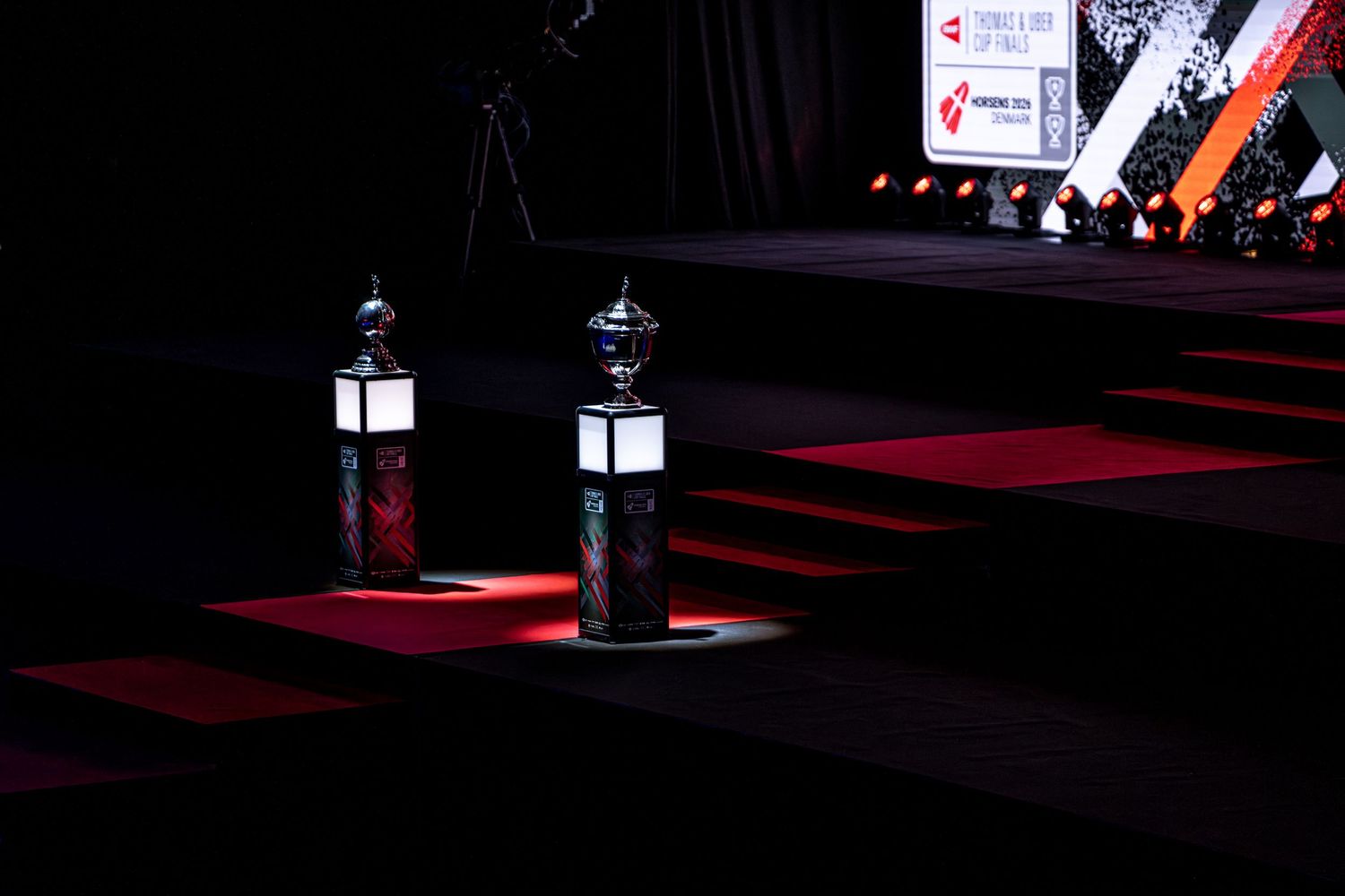 Two ornate trophies spotlit on pedestals on a dark stage at a badminton championship.