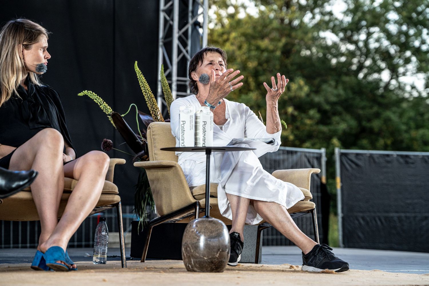 Woman in white outfit gesturing expressively during an outdoor panel discussion on stage.