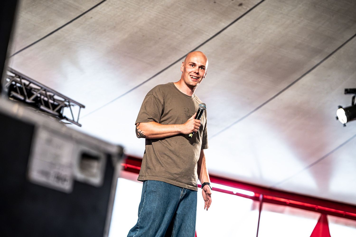 Bald man in olive t-shirt and jeans speaking on outdoor stage holding microphone.