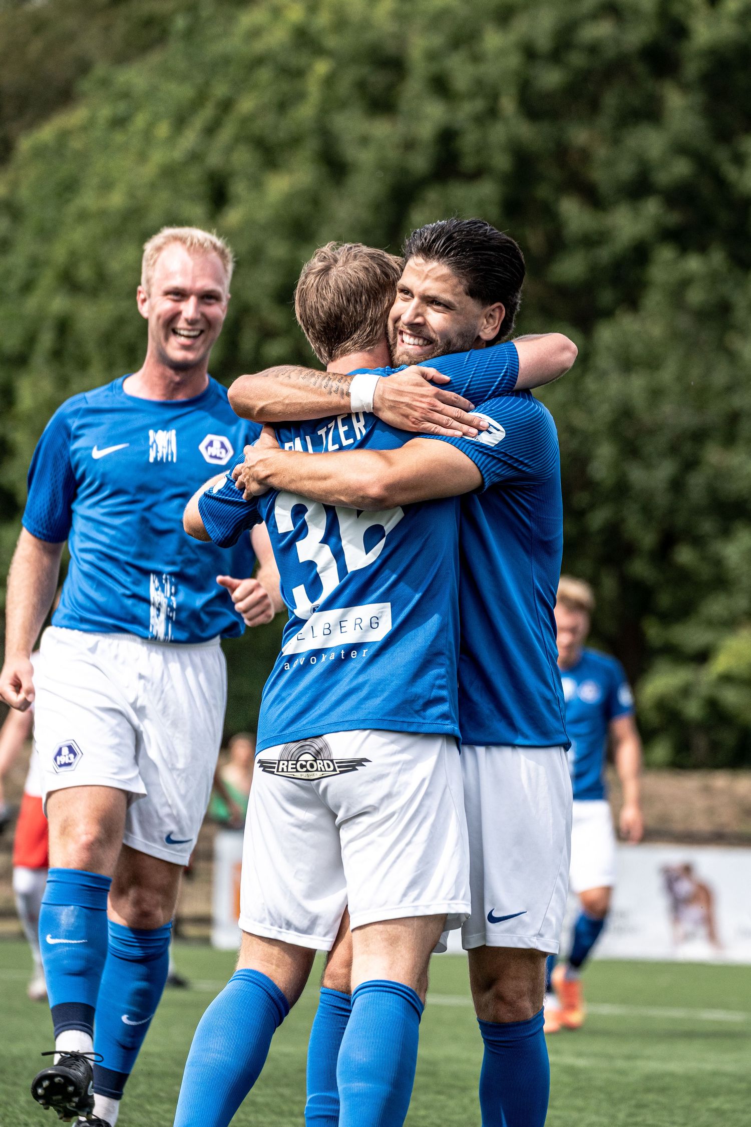 Soccer players in blue and white Nike uniforms share a celebratory hug on a sunny outdoor pitch.