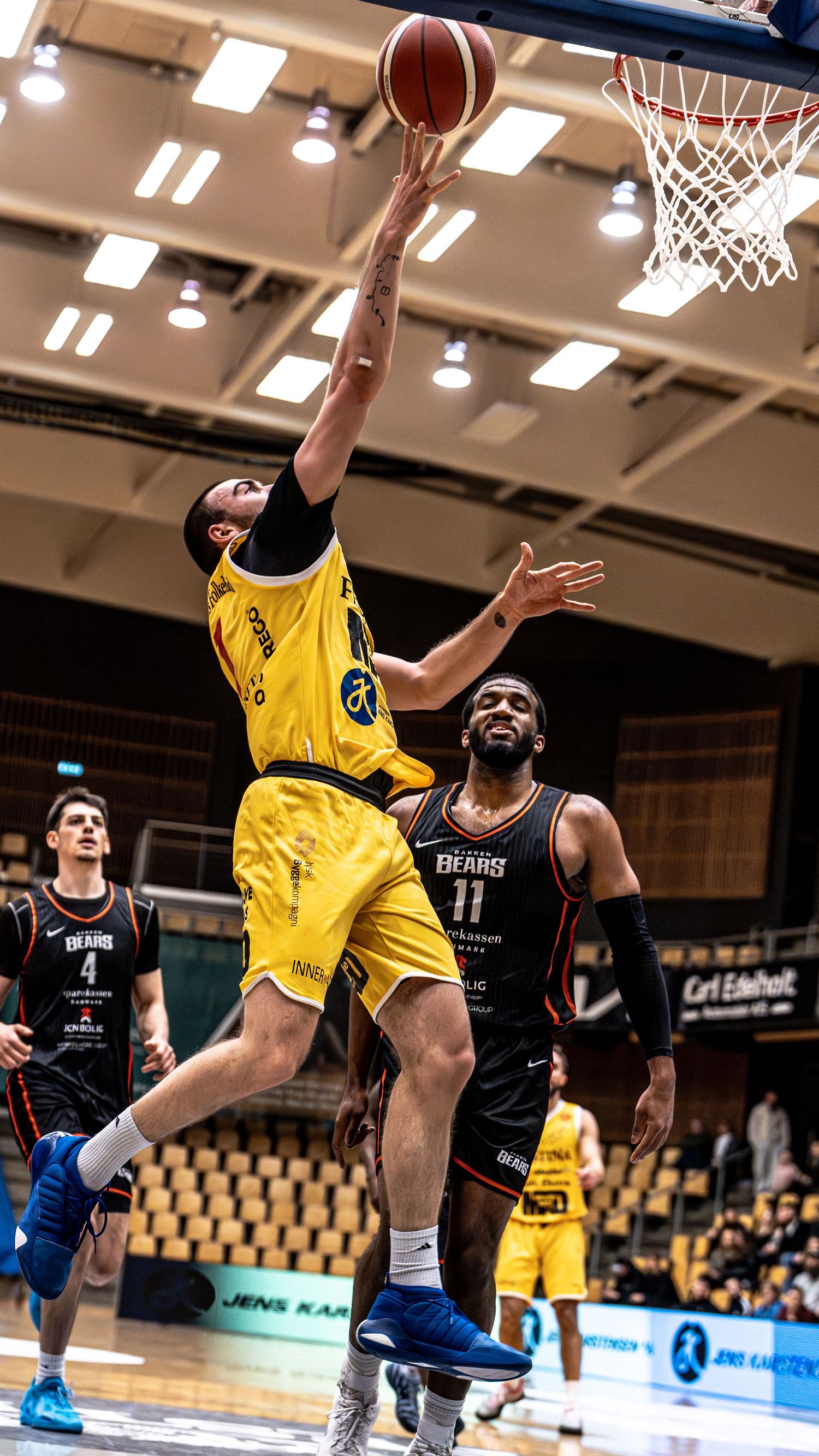 Yellow jersey player leaps for a layup while defender in Bears jersey watches.