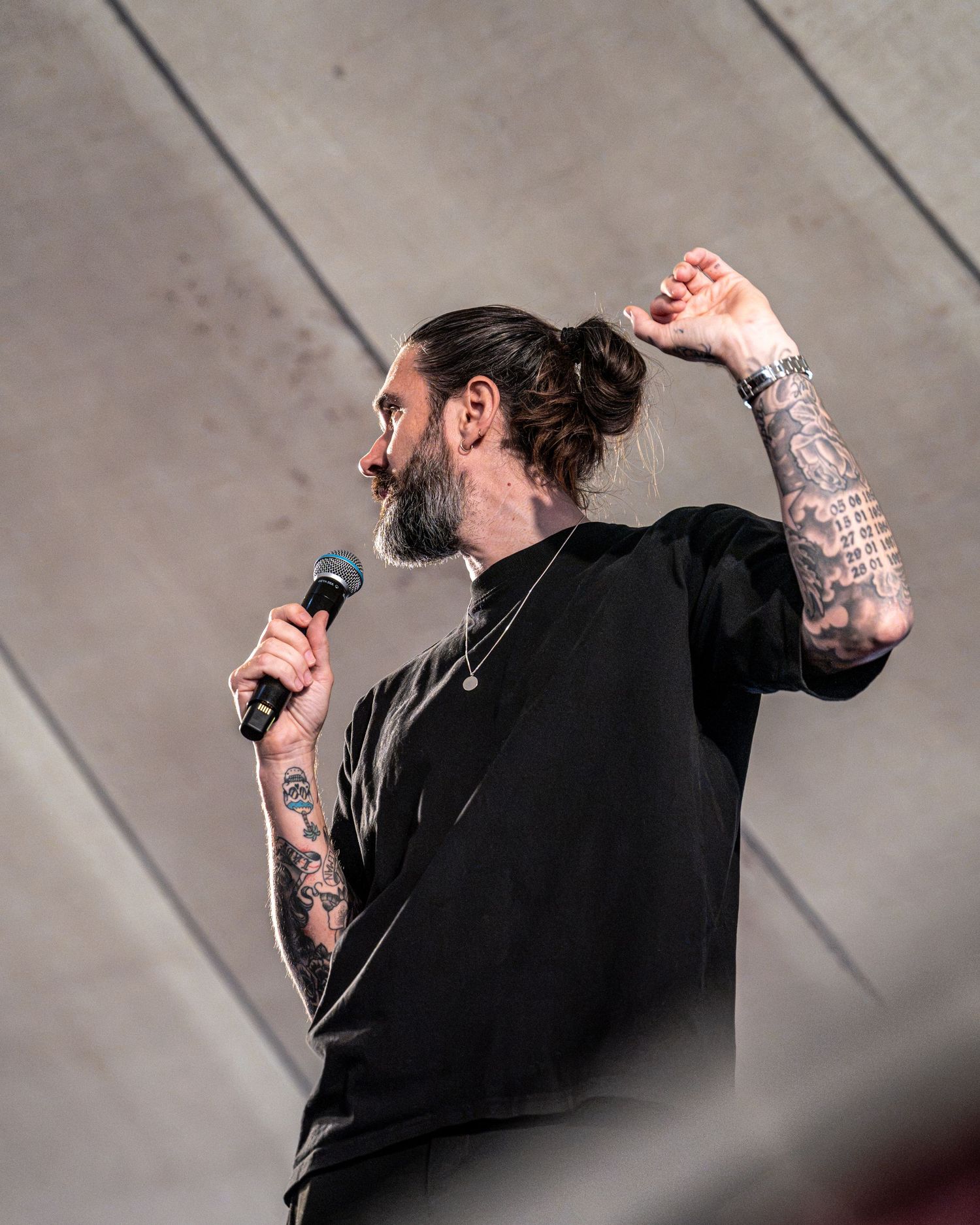 Tattooed bearded performer in black outfit holding microphone while gesturing with raised hand on stage.
