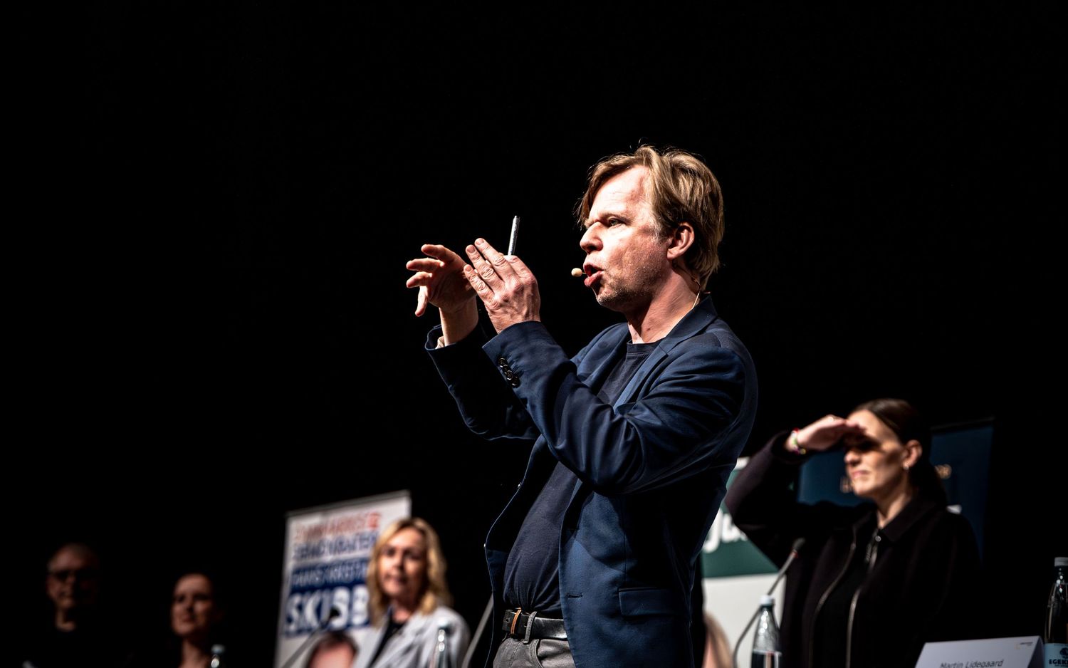 Man in navy blazer gesturing on a dark stage during a live performance or speaking event.