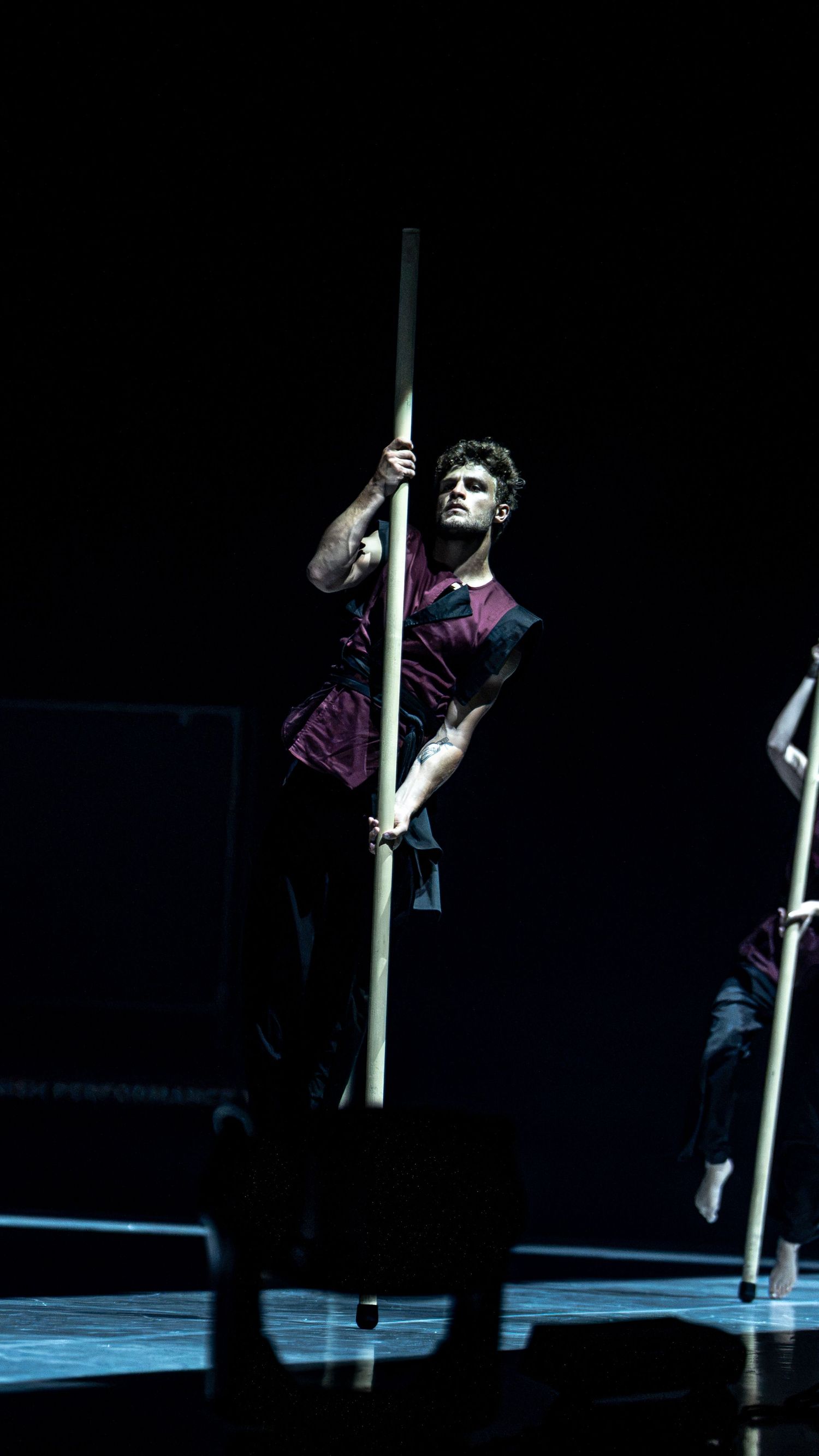 Male dancer in burgundy costume holds tall pole on dark stage during theatrical performance.