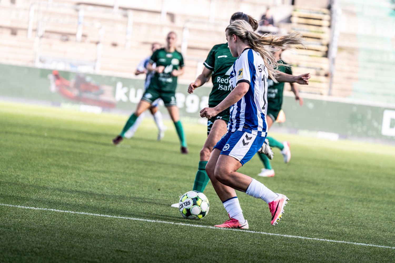 Female footballer in blue and white strips drives forward with the ball in a women's league soccer game.