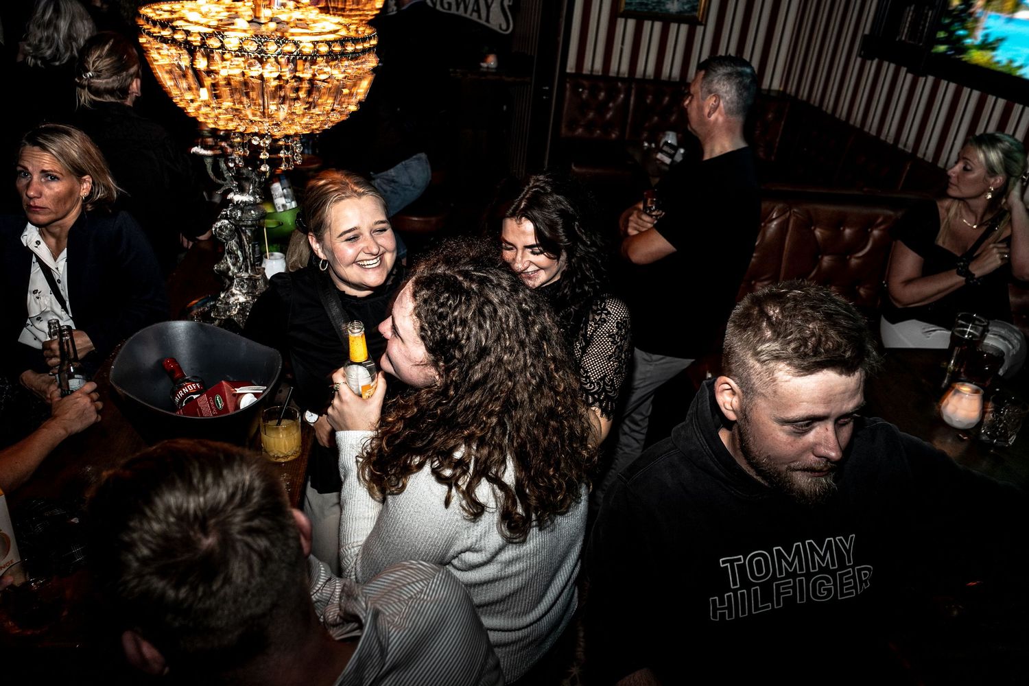 Group of friends laughing and chatting around a table in a cozy, dimly lit bar with a warm chandelier overhead.