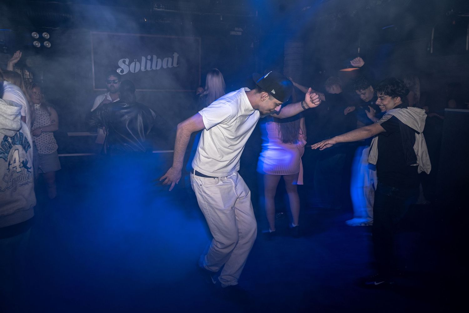 Man in white shirt and pants dances energetically in a smoky, blue-lit nightclub as other partygoers dance in the background.