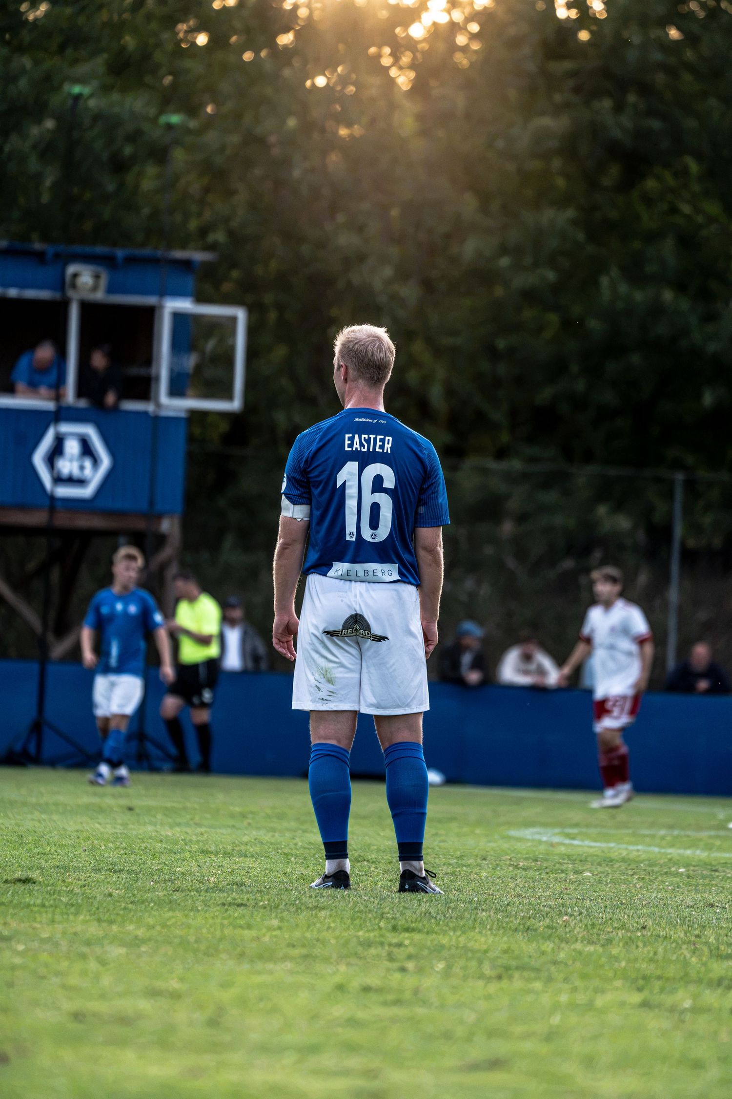 Soccer player Easter number 16 in blue kit stands on pitch during a sunset match.