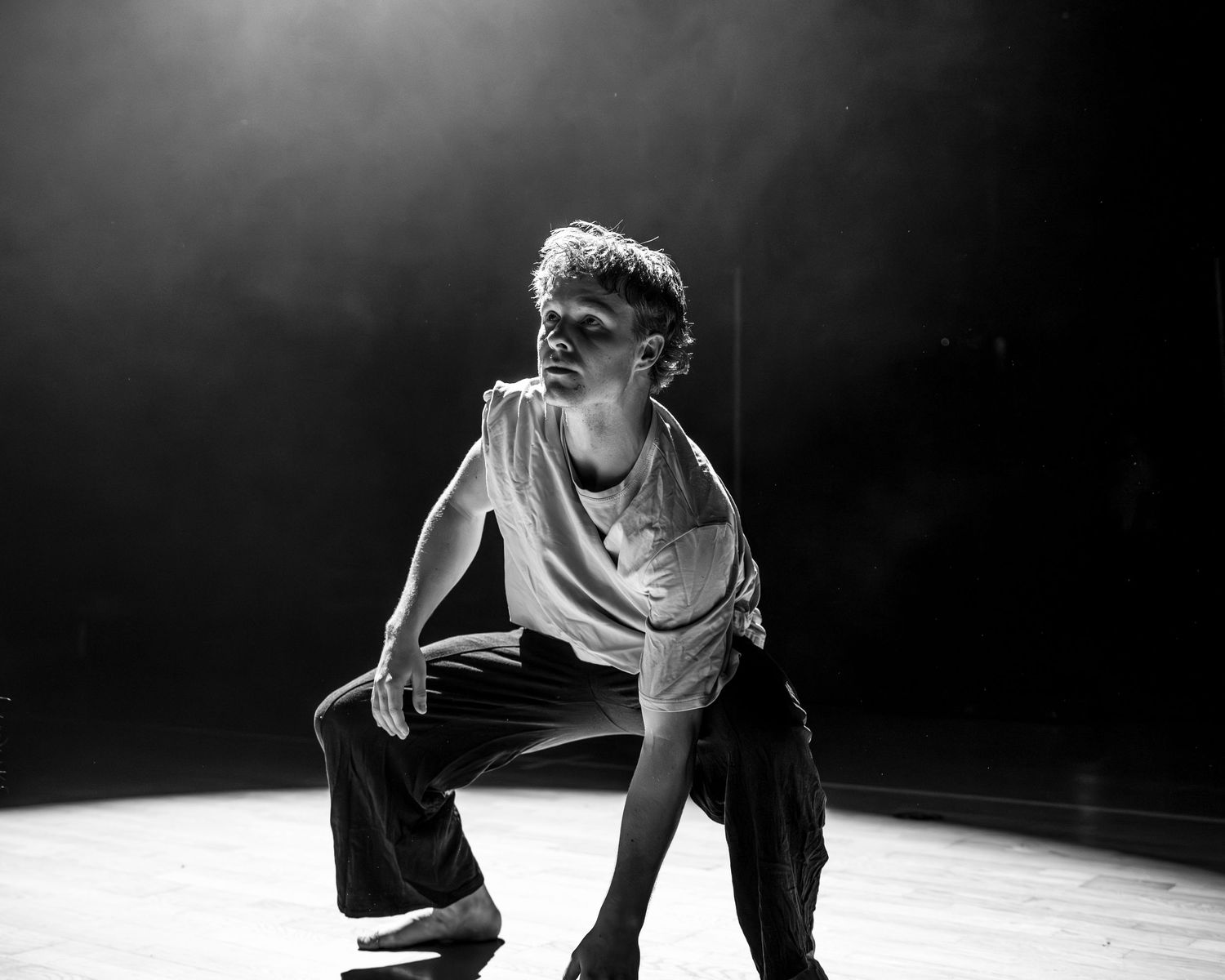 Male dancer crouches dramatically on stage floor in black and white performance photo.