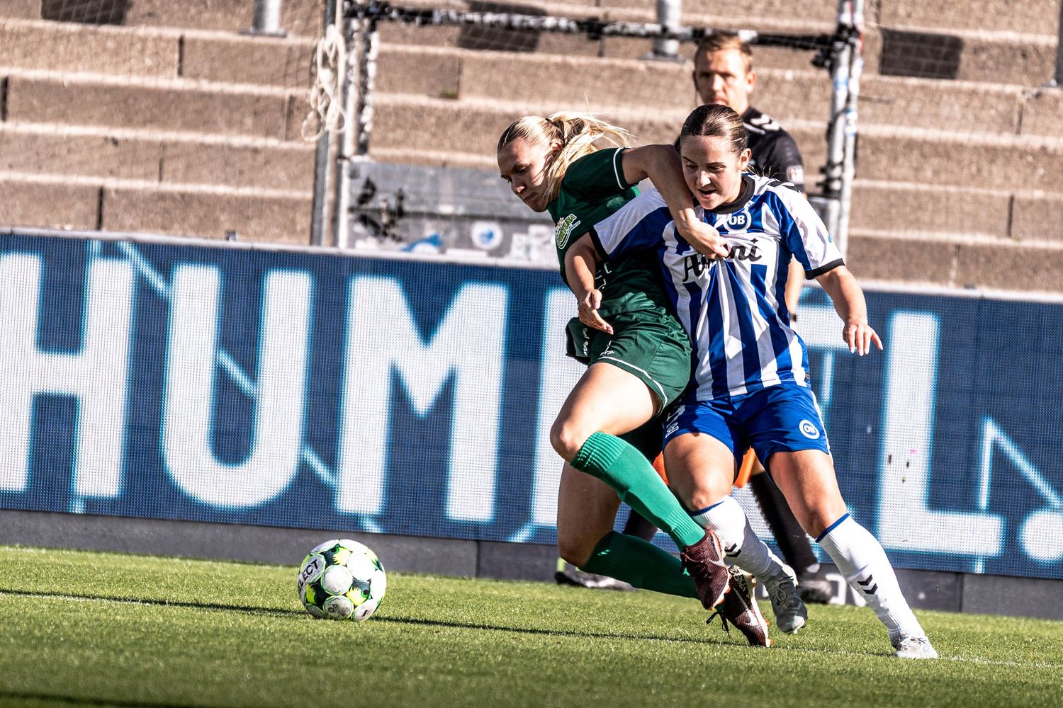 Two female soccer players competing for the ball, one in green and one in blue and white, during a match.