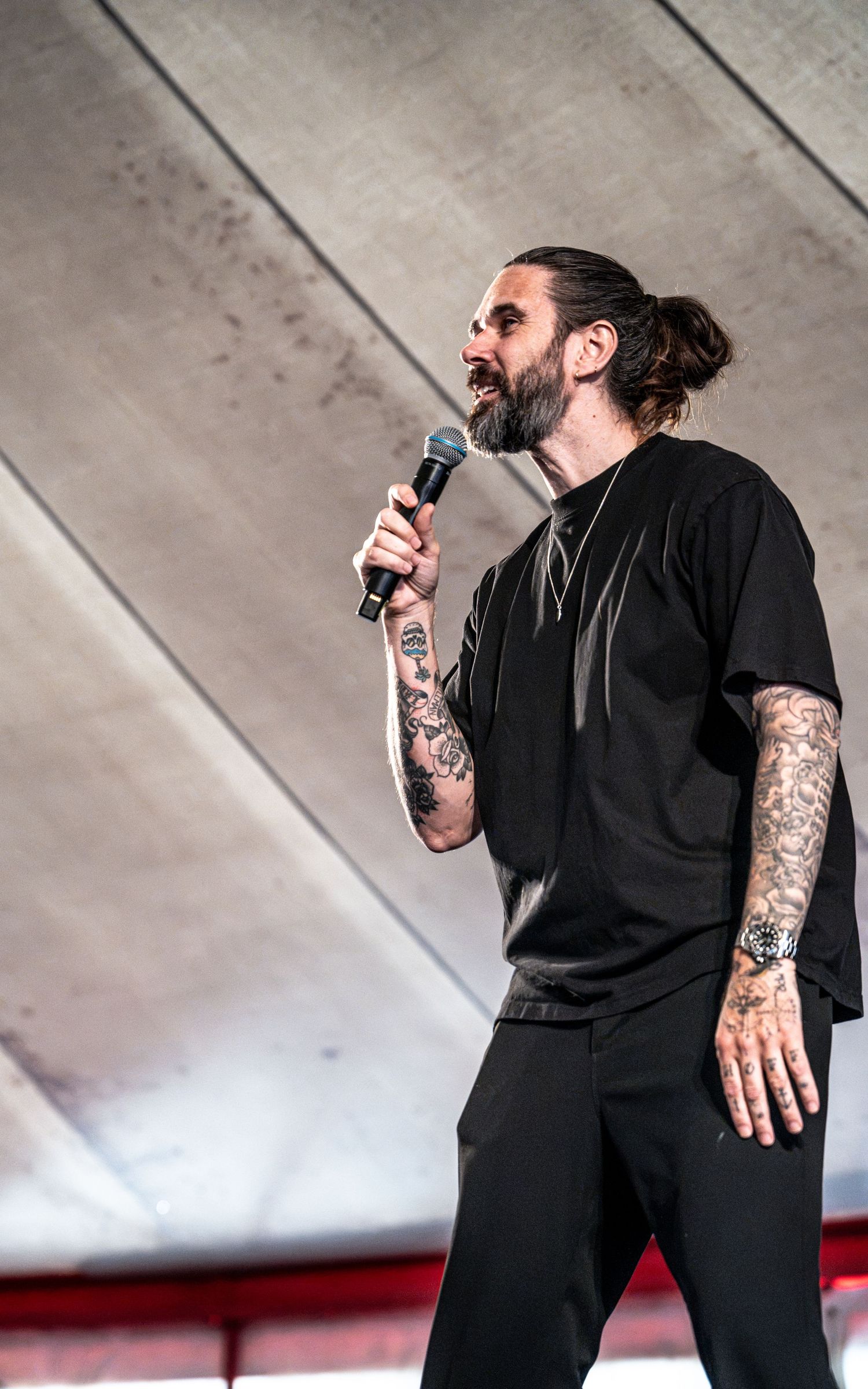 Bearded tattooed man in black outfit singing passionately into microphone on outdoor stage.