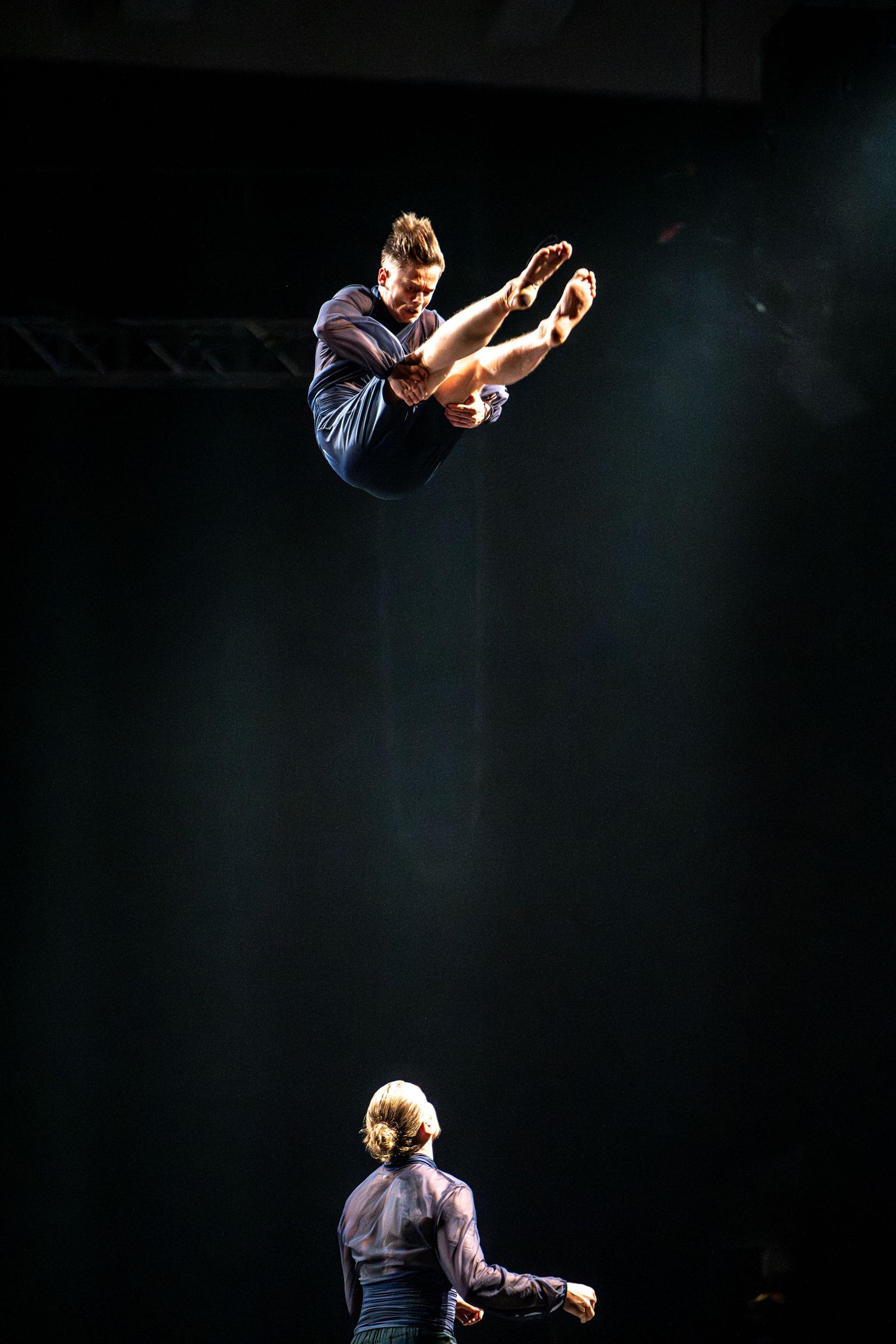 Acrobat soaring high above another performer on a dark stage during a live circus performance.