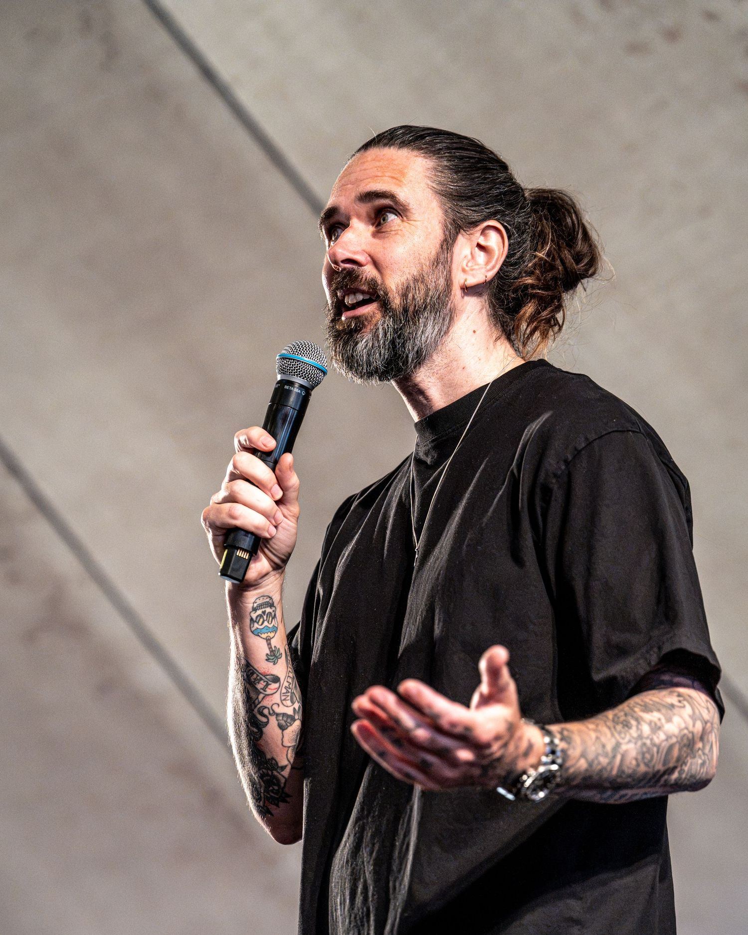 Bearded tattooed man in black t-shirt speaking into blue microphone on outdoor stage.