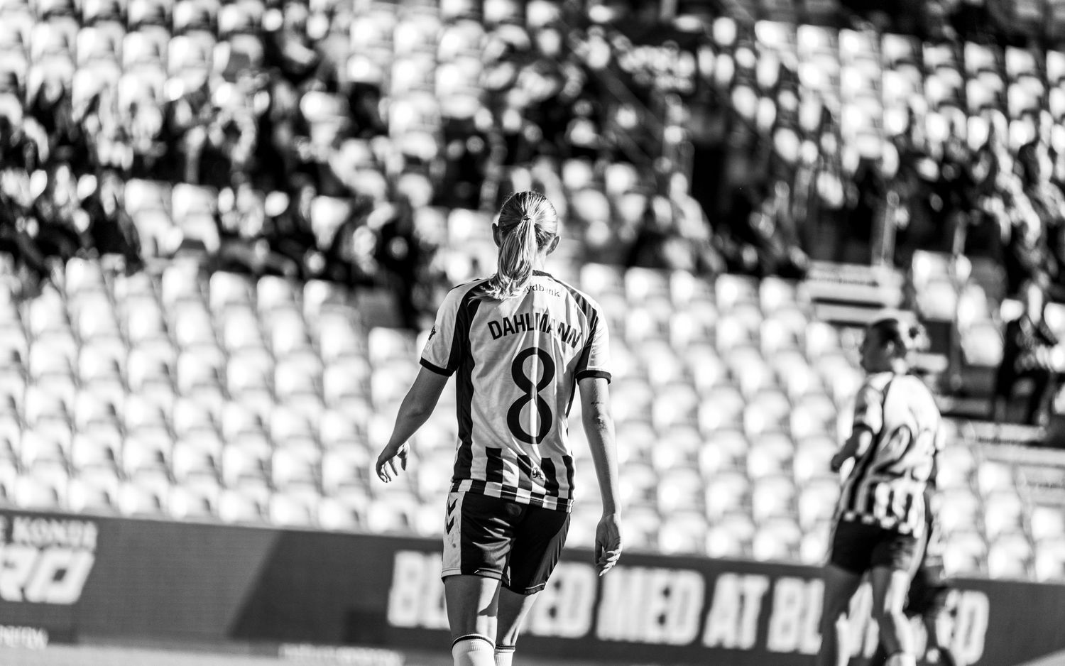Black and white photo of female soccer player number 8 standing on pitch with stadium crowd in background.