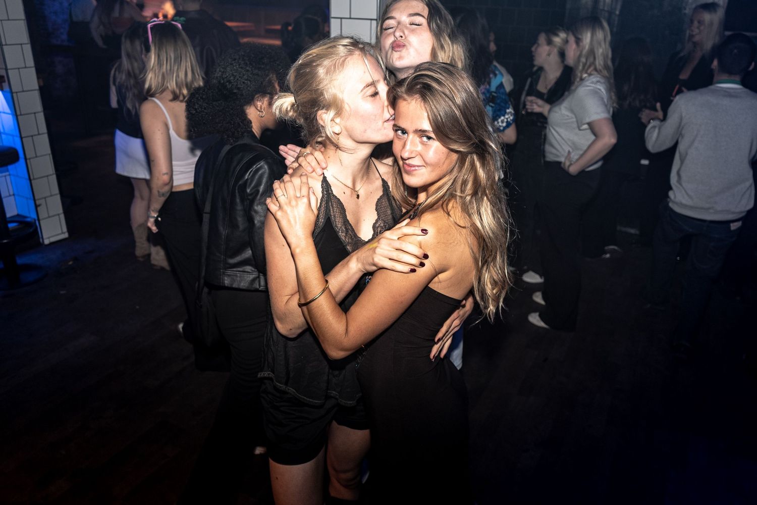 Two young women with long blonde hair embrace and pose closely together on a crowded nightclub dance floor with friends behind them.