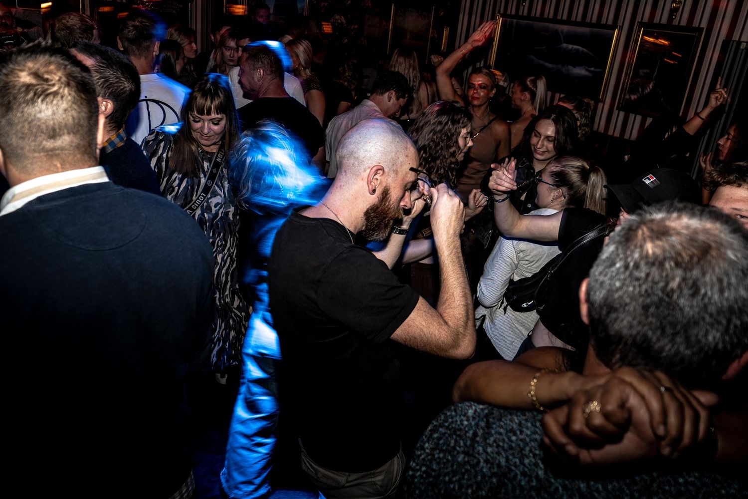 A bald man in a black t-shirt dances in a crowded, dimly lit bar as blue light glows around the lively nightlife crowd.