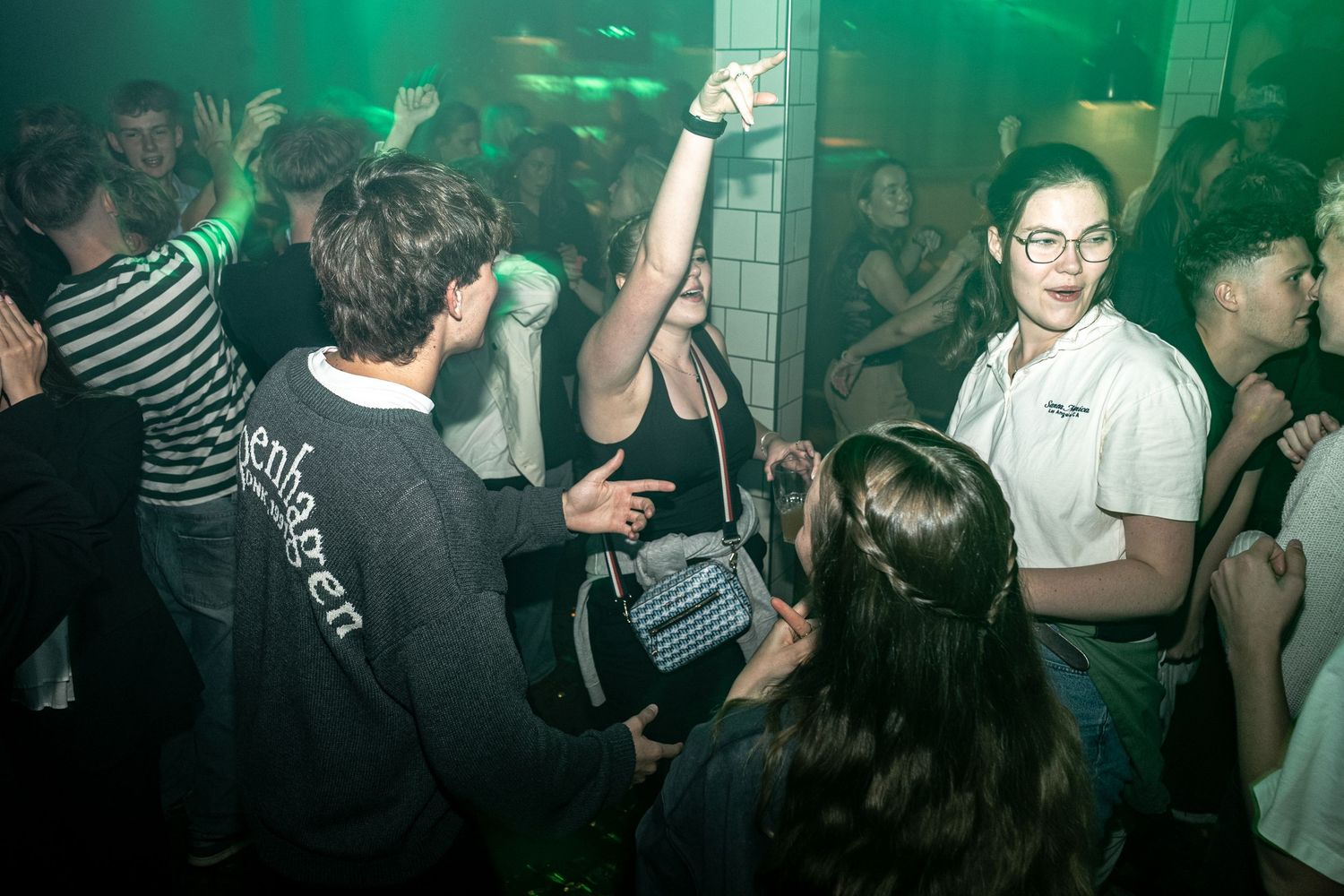 Crowd dancing at a nightclub party with green lighting, a woman in glasses smiling and a person raising an arm in the air.