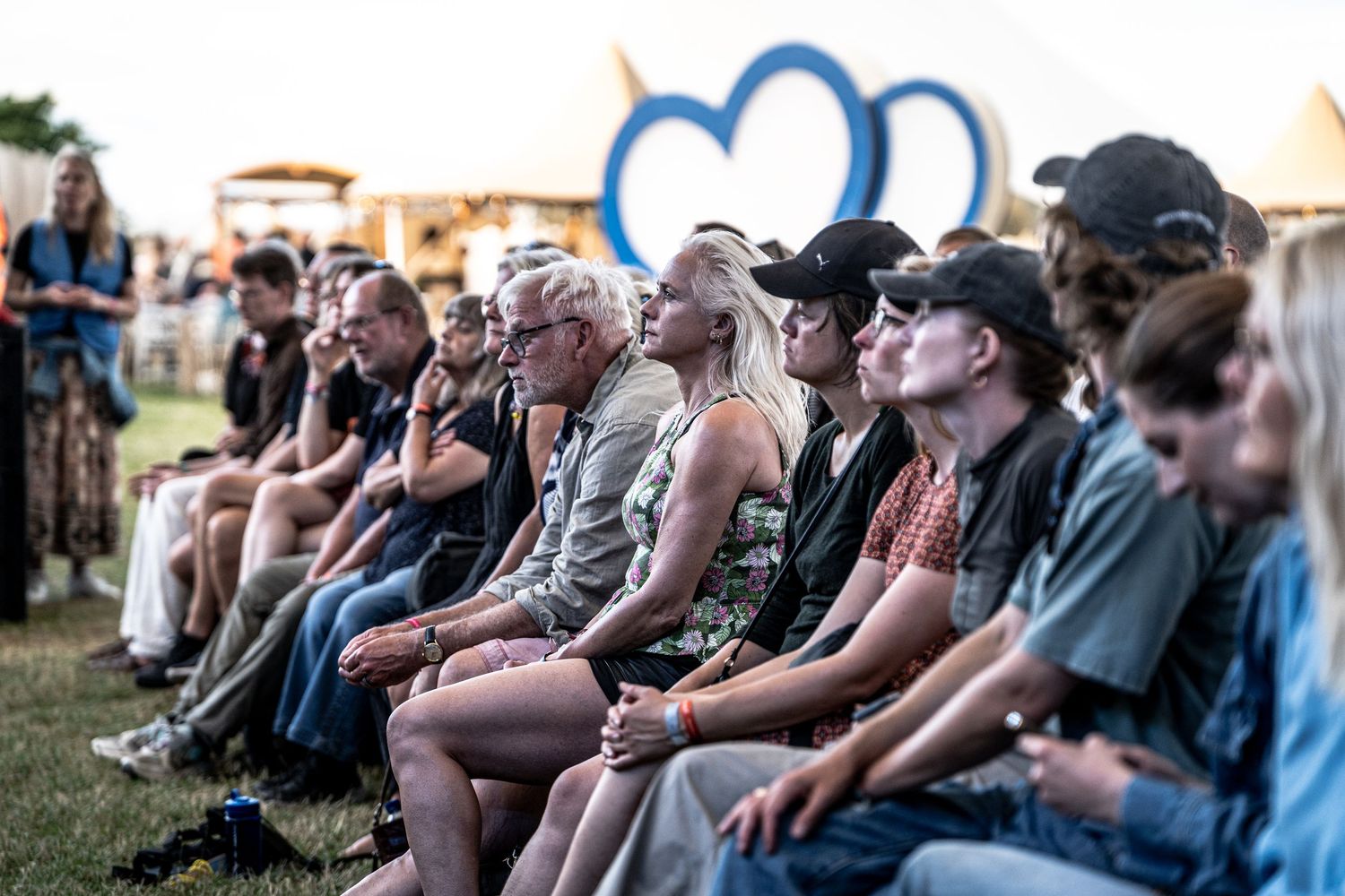 Crowd of festival attendees sitting on grass in front of large blue heart sculpture at outdoor event.