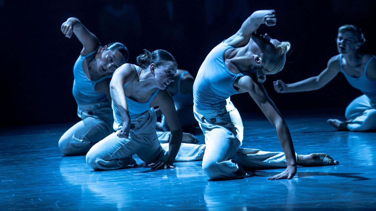Contemporary dance performers in white costumes moving expressively on a blue-lit stage during a theatrical production.
