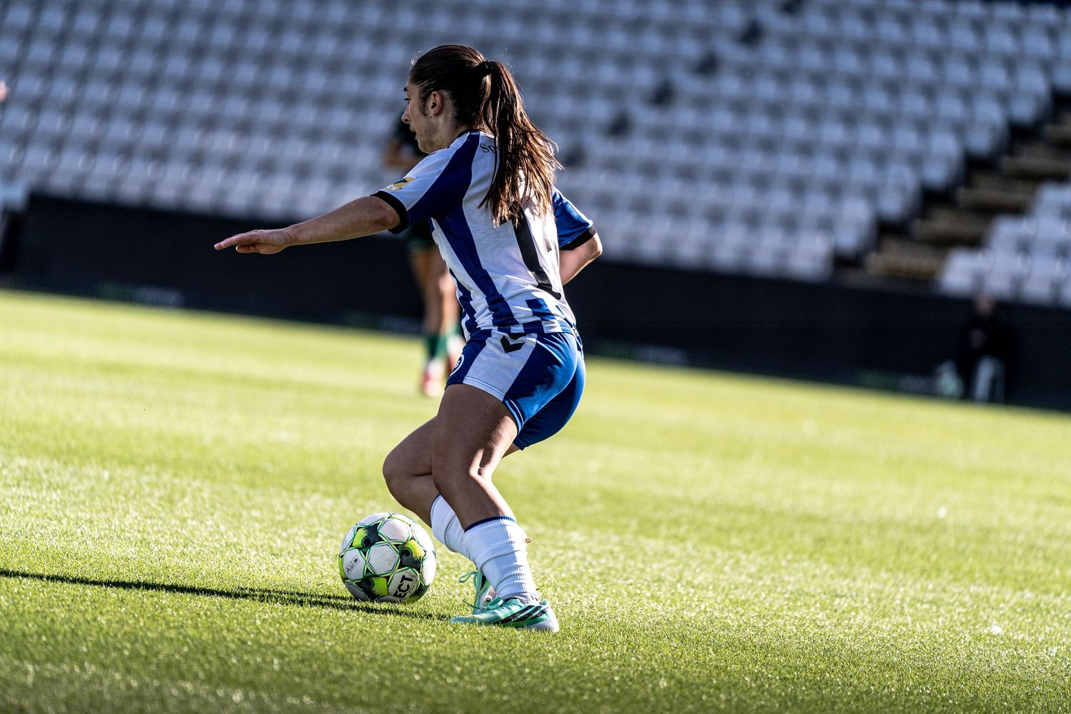 Female soccer player in blue and white striped kit dribbles ball on grass pitch in empty stadium.