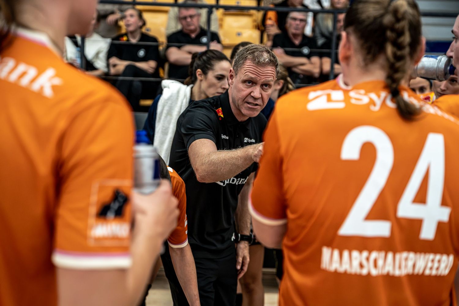 Male handball coach in black outfit giving tactical instructions to female players in orange jerseys.