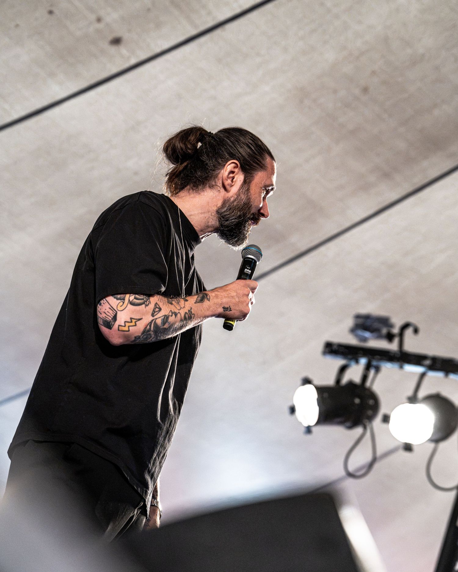 Side view of tattooed male vocalist with man bun singing into microphone on stage.