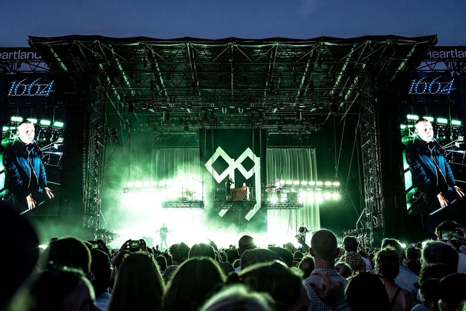 Large outdoor concert stage with green lights and crowd at night music festival.
